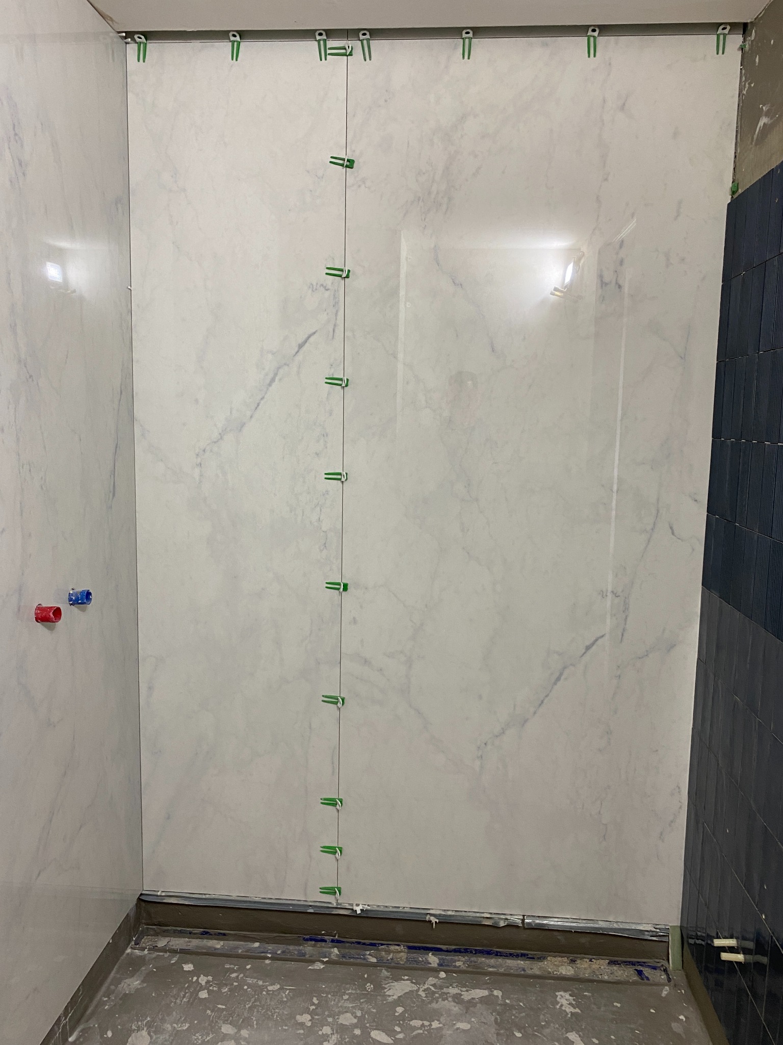 Ściana łazienki w trakcie układania płytek marmuropodobnych, z widocznymi klipsami poziomującymi i krzyżykami dystansowymi, fragment niebieskich płytek po prawej stronie.