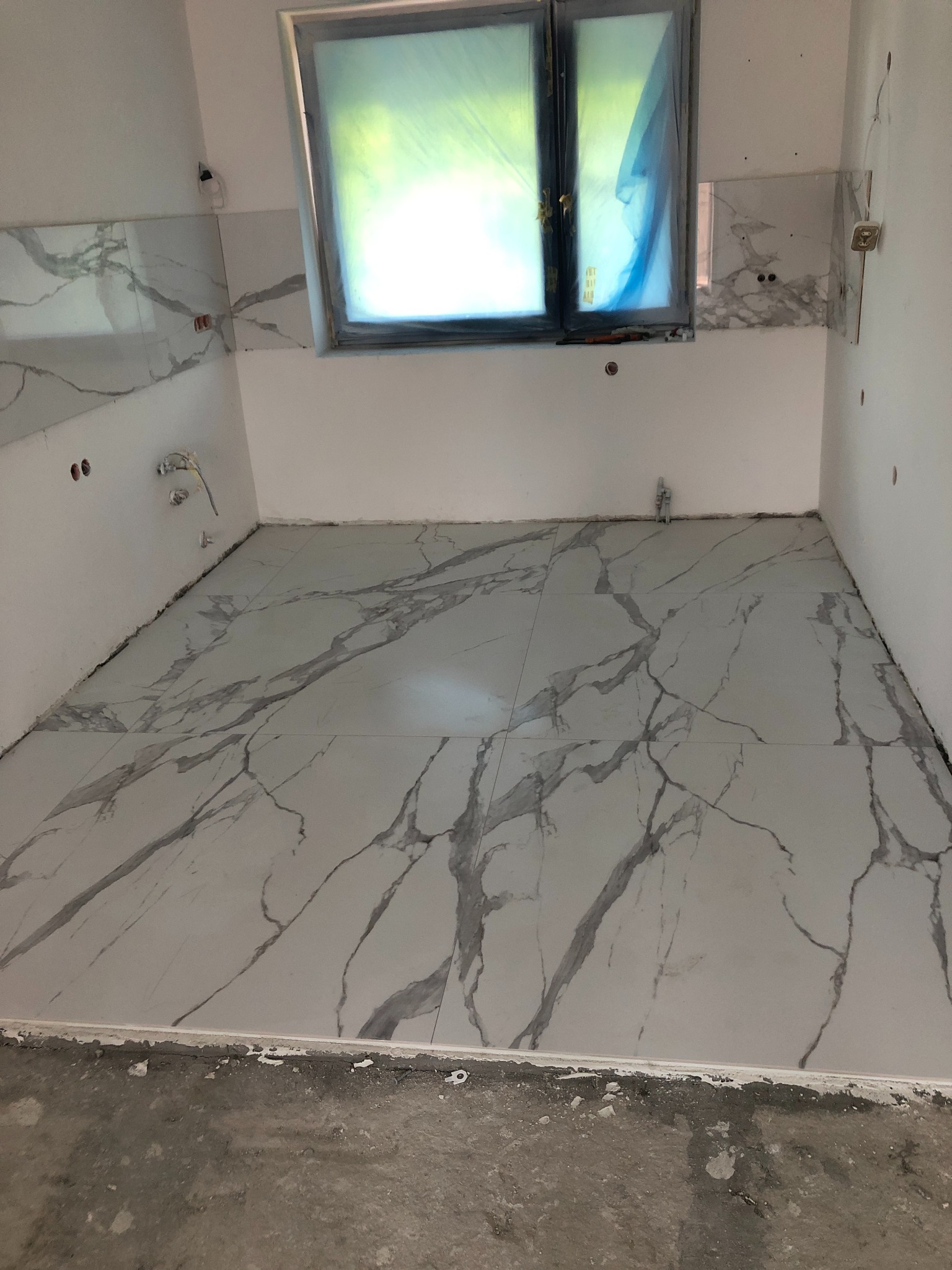 Łazienka w trakcie prac: położone płytki podłogowe i częściowo ścienne z motywem białego marmuru z szarymi żyłami, okno zabezpieczone folią, widoczne niedokończone instalacje elektryczne.