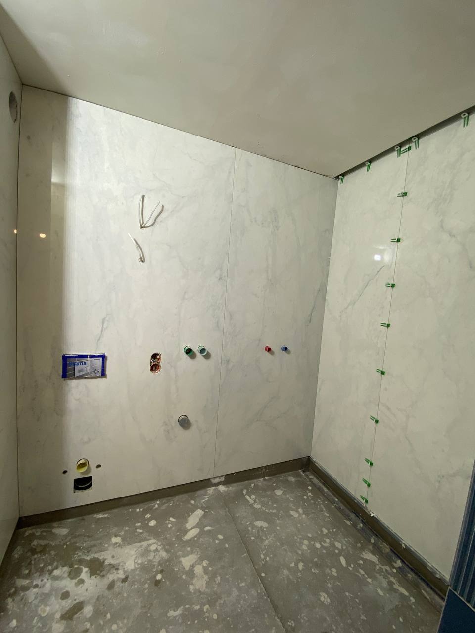 Łazienka w trakcie wykańczania: białe płytki imitujące marmur na ścianach, widoczne otwory na instalacje, zielone krzyżyki dystansowe między płytkami, surowa betonowa podłoga z resztkami zaprawy.