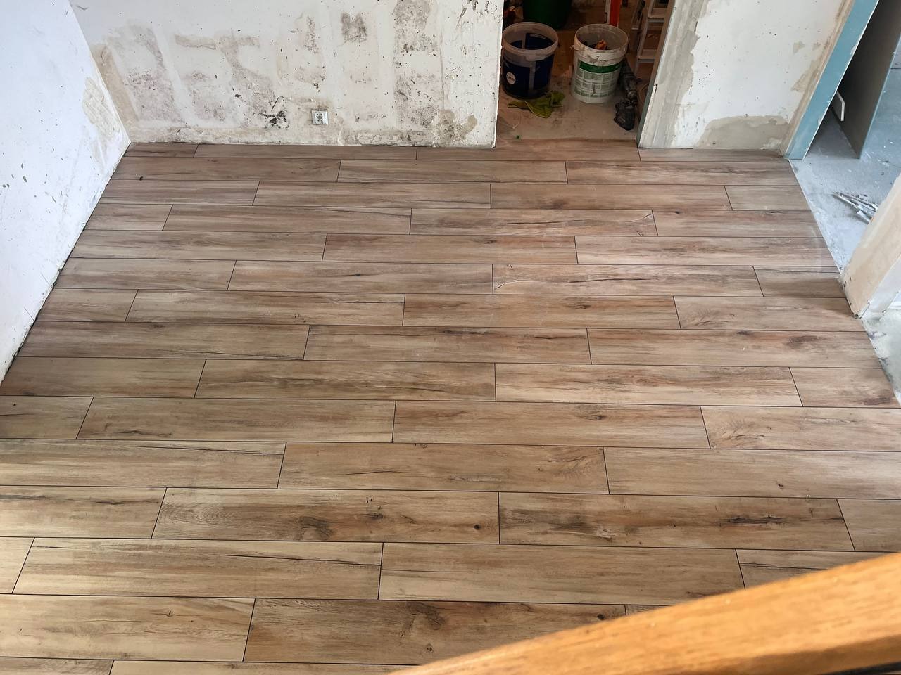 Ułożone płytki podłogowe imitujące drewno w trakcie remontu łazienki, widoczne ślady po kleju na ścianach i wiaderka z zaprawą w tle.