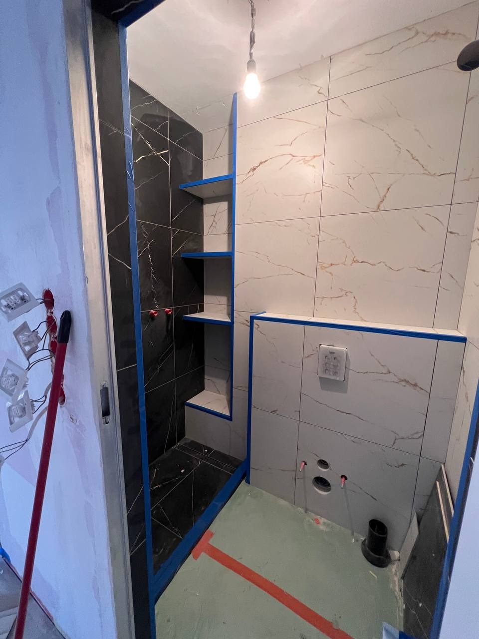 Remontowana łazienka: czarne i białe płytki imitujące marmur, półki w kabinie prysznicowej, widoczne instalacje hydrauliczne przed montażem armatury, zabezpieczenia taśmą malarską.