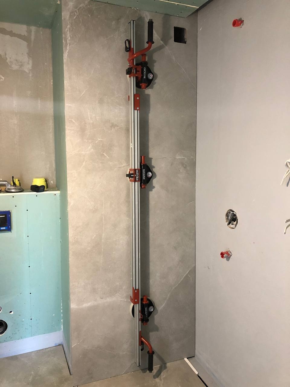 Ustawiony pionowo na ścianie w łazience przyrząd do cięcia płytek ceramicznych z trzema przyssawkami, widoczne niedokończone ściany i instalacje.