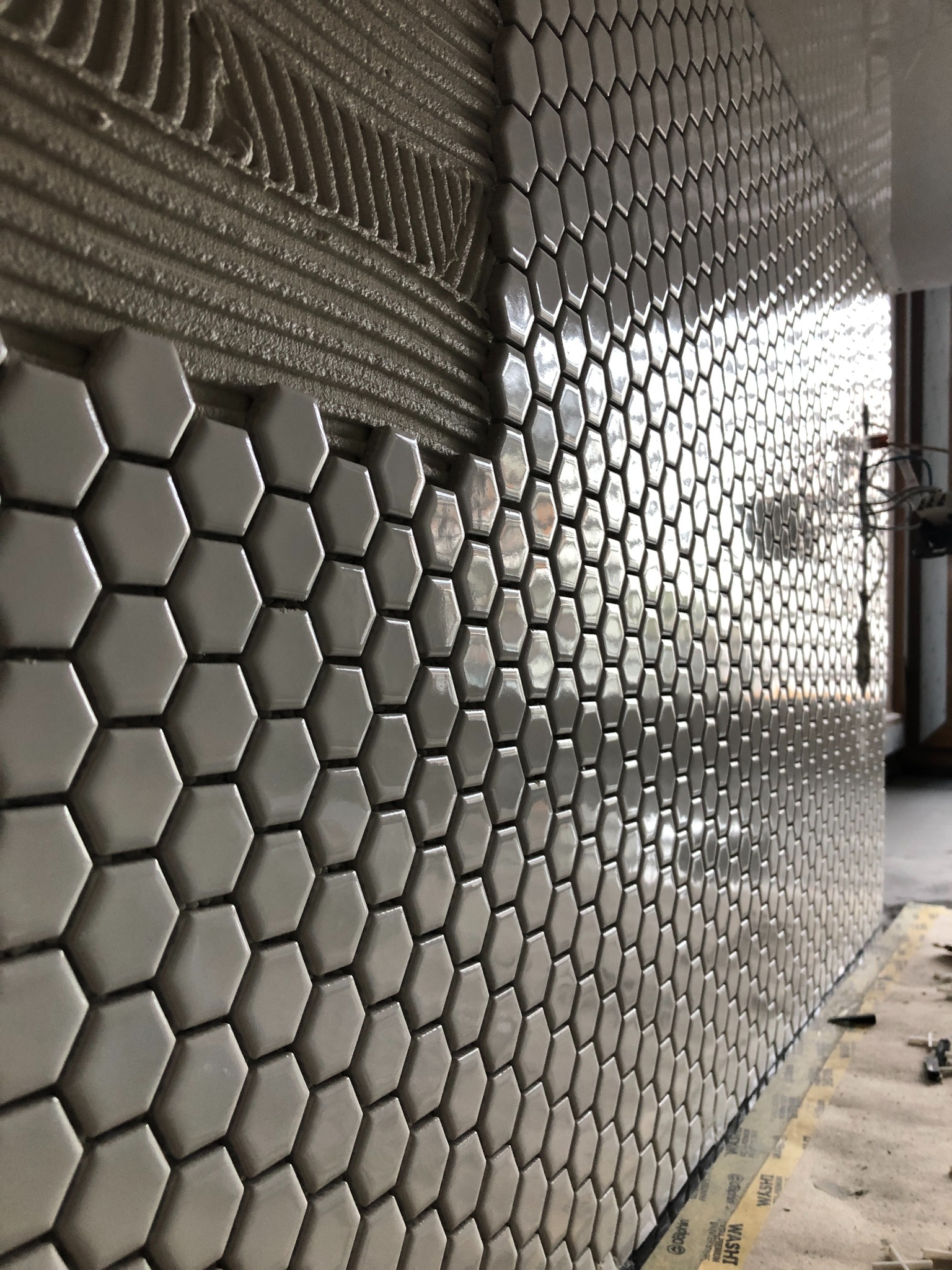 Układanie heksagonalnych, szarych płytek na ścianie, widoczna warstwa kleju i ułożone kafelki, zbliżenie na etap prac.