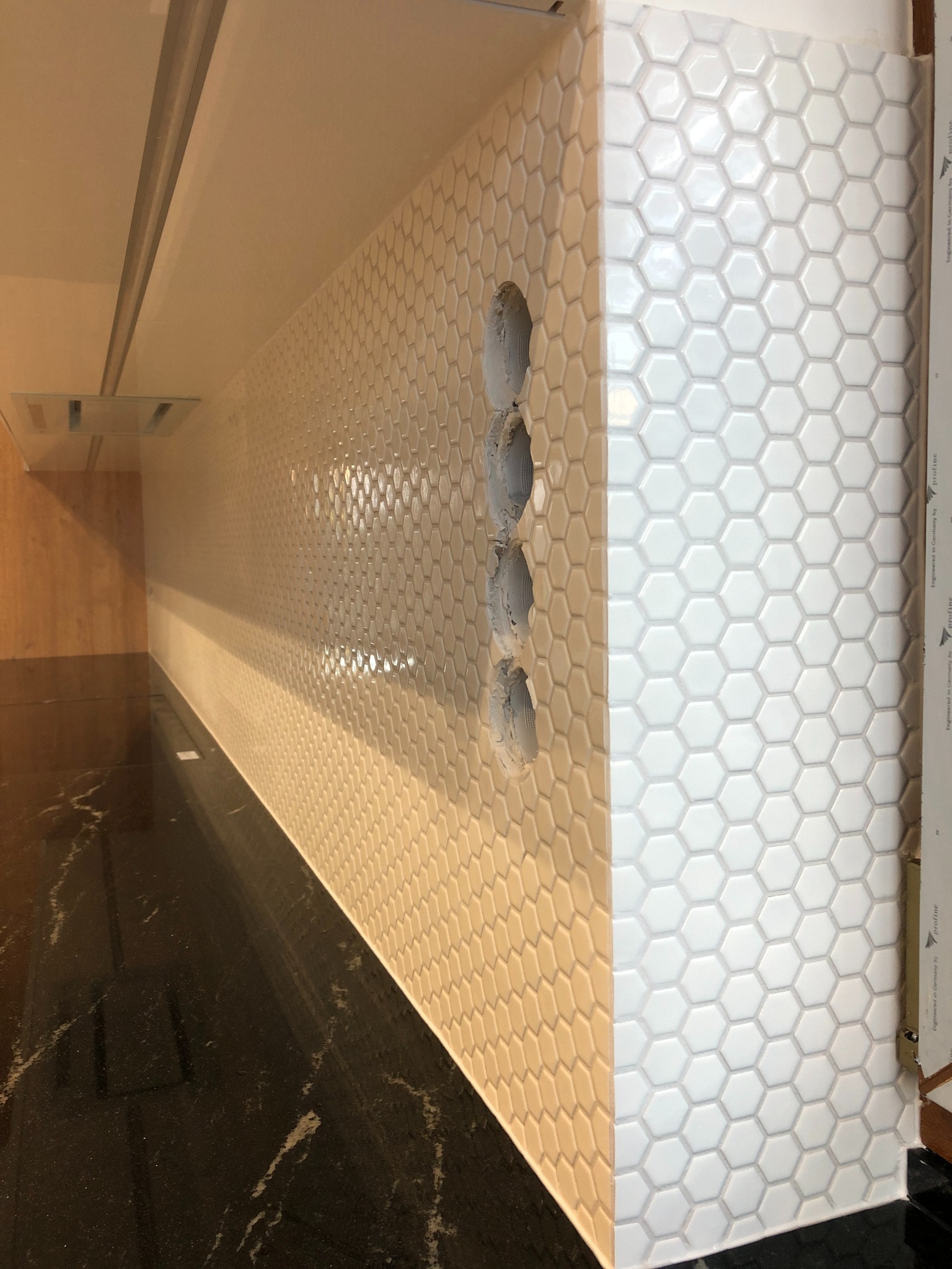 Ściana wyłożona białymi, heksagonalnymi płytkami z widocznymi otworami na gniazdka elektryczne, częściowo wypełnionymi szarą zaprawą. Czarny blat z marmurkowym wzorem w dolnej części kadru.