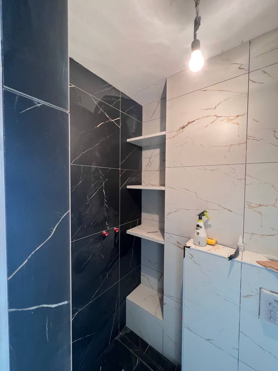 Częściowo wykończona łazienka z czarnymi i białymi marmurkowymi płytkami, z widocznymi półkami wnękowymi i narzędziami glazurniczymi na blacie.