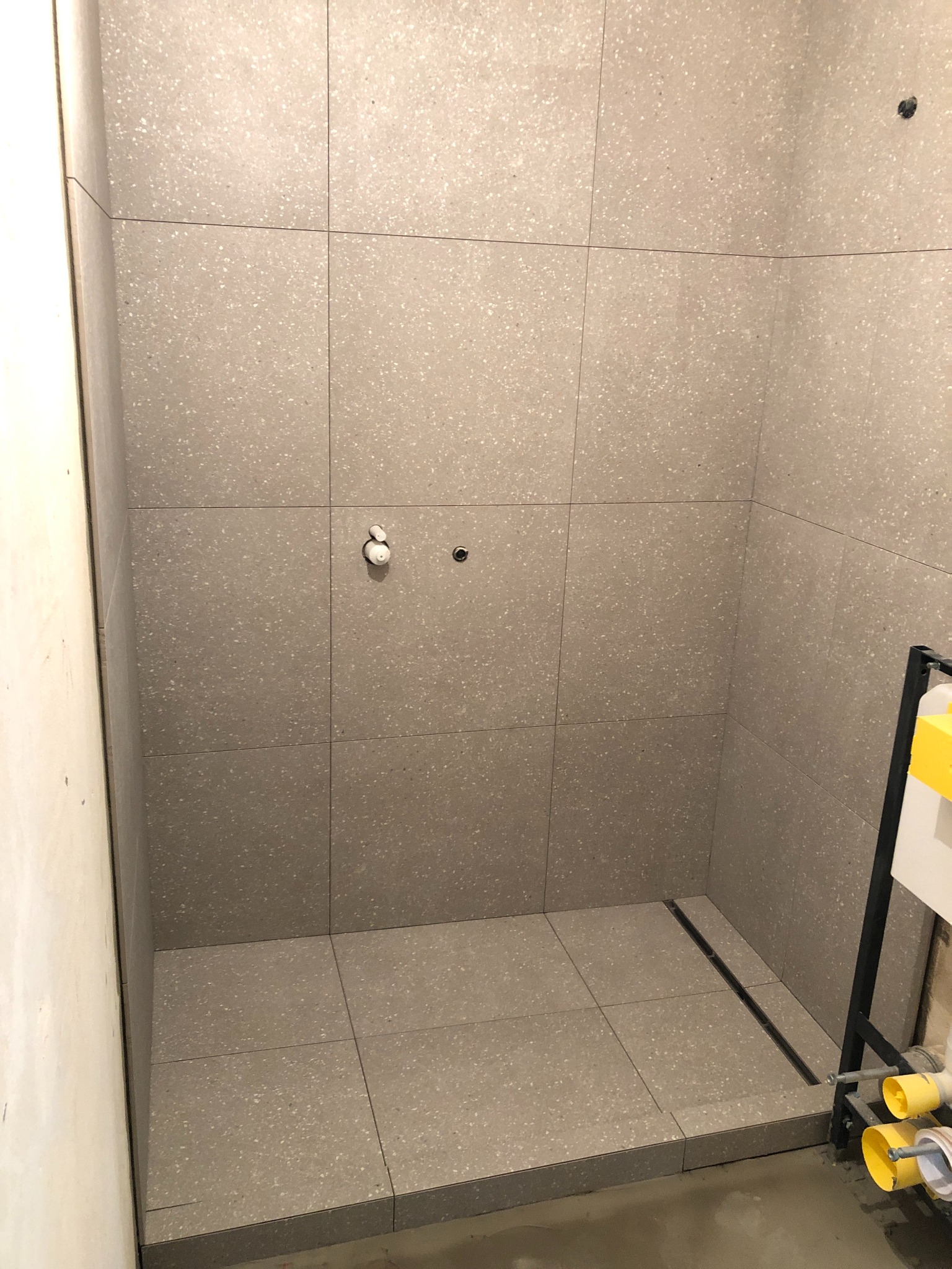 Prysznic w trakcie wykańczania, szare płytki imitujące lastryko na ścianach i podłodze, widoczny odpływ liniowy i instalacja wodna bez armatury, surowa ściana obok.
