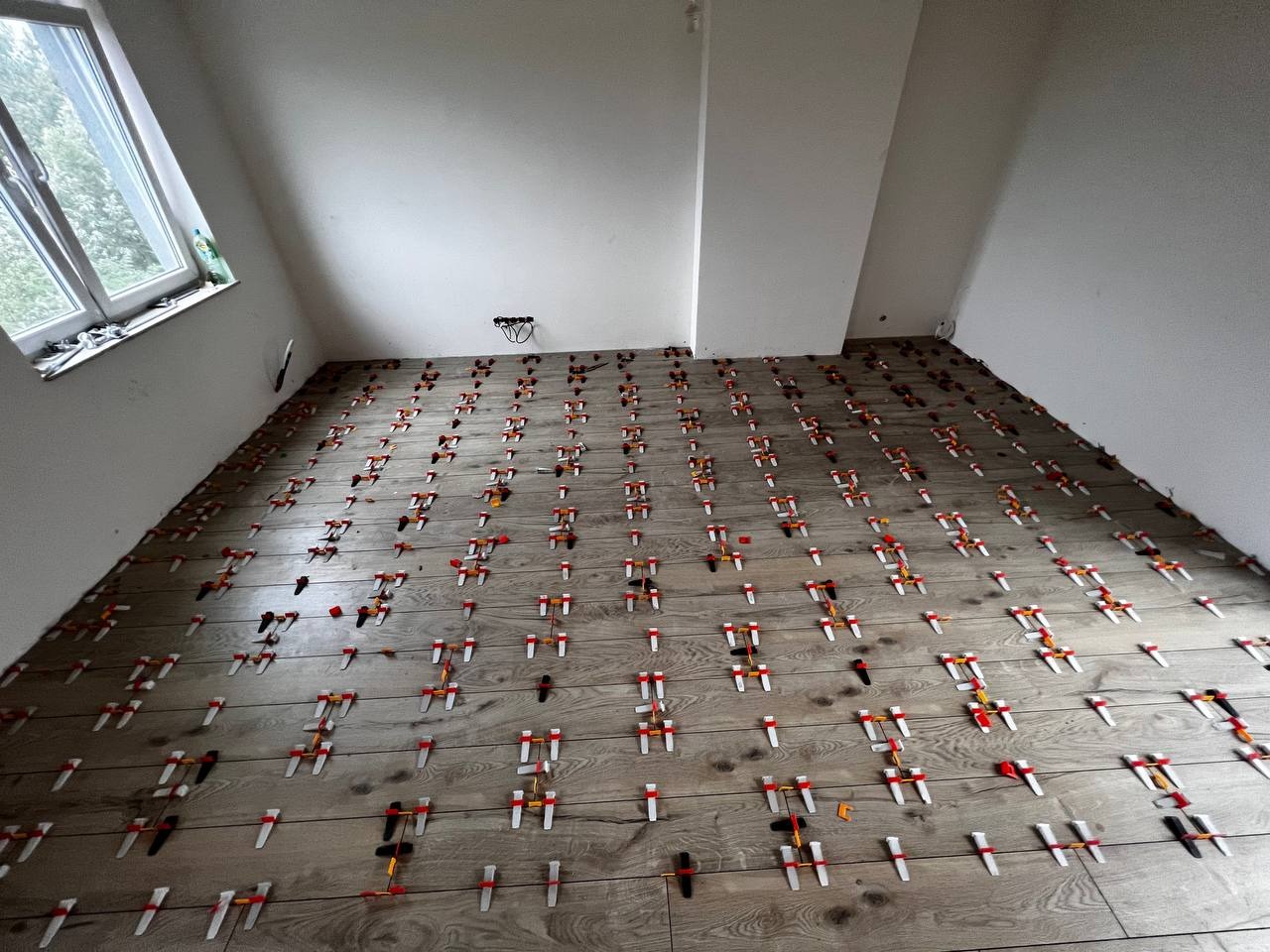 Układanie płytek podłogowych imitujących drewno z użyciem systemu poziomowania, widoczne kliny i klipsy na całej powierzchni, surowe ściany w tle.