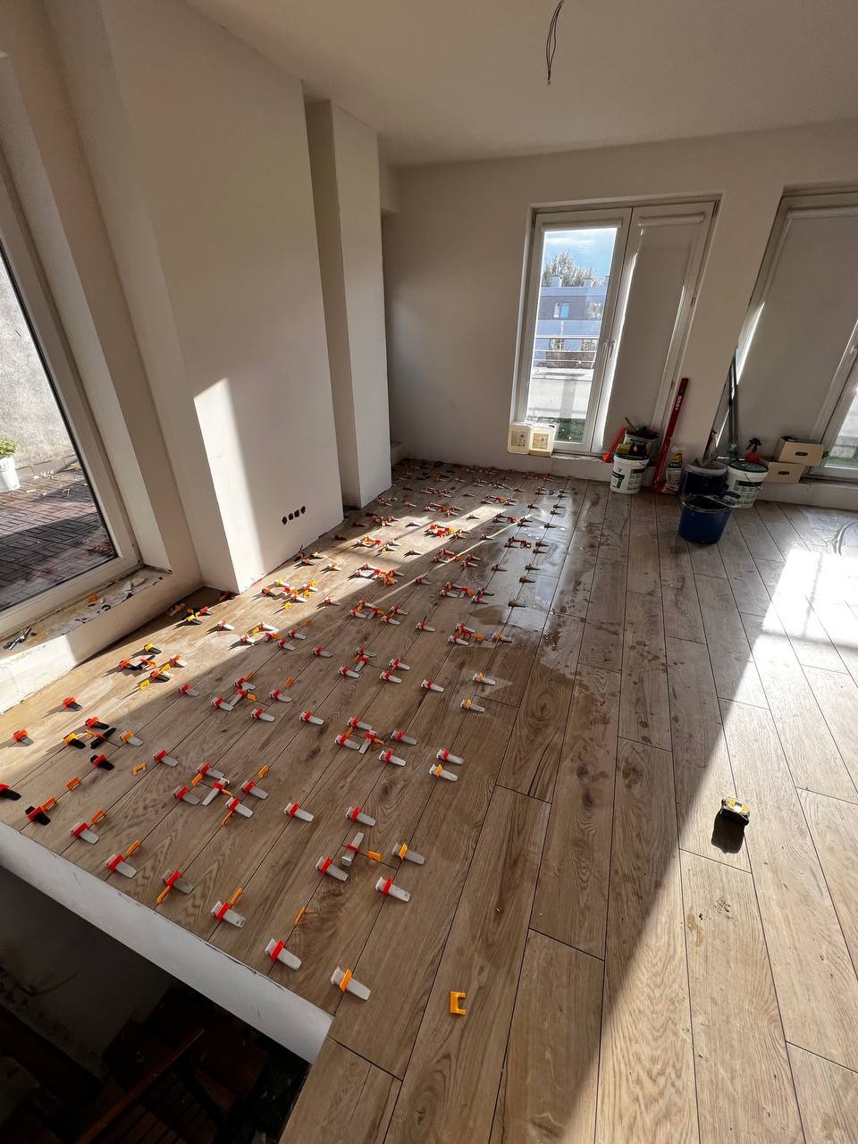 Układanie płytek imitujących drewno w jasnym pomieszczeniu z dużymi oknami, widoczne klipsy poziomujące i narzędzia glazurnicze.