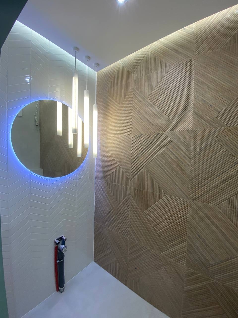 Nowoczesna łazienka z drewnopodobną mozaiką na ścianie, owalnym lustrem z niebieskim podświetleniem LED, białymi płytkami ułożonymi w jodełkę i minimalistycznymi lampami wiszącymi.