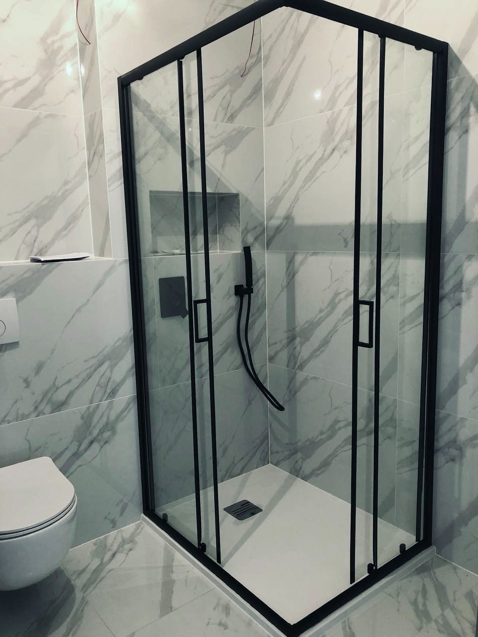 Kabina prysznicowa z czarnymi profilami na tle marmurowych płytek, widoczny odpływ liniowy i toaleta.