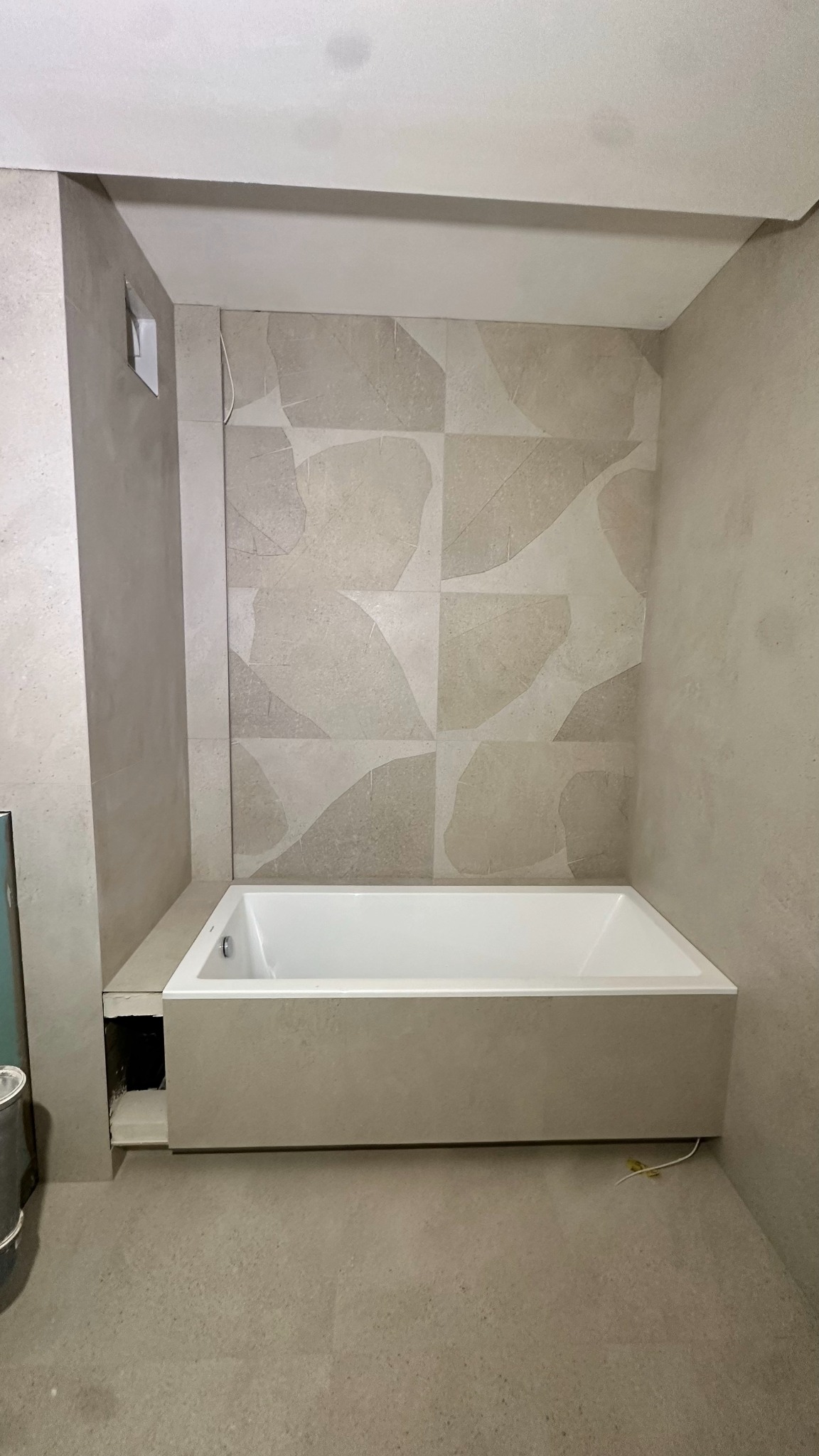 Wanna obudowana płytkami w łazience z beżowymi płytkami z motywem liści. Widoczne niedokończone prace przy obudowie wanny i wystające kable.