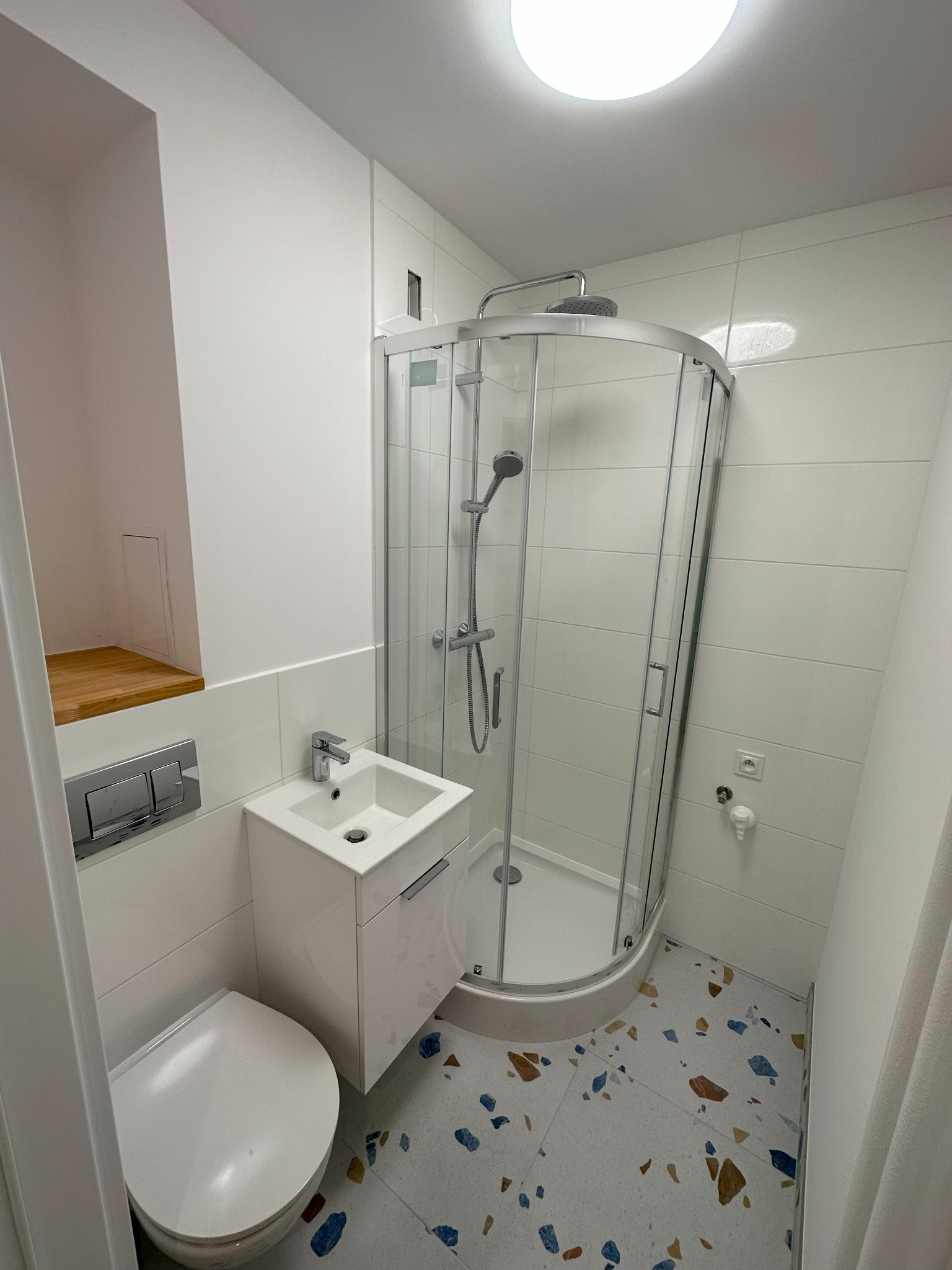 Nowoczesna, mała łazienka z kabiną prysznicową z zaokrąglonym szkłem, wiszącą umywalką z szafką, podwieszanym WC i podłogą w stylu lastryko, oświetlona okrągłym plafonem.