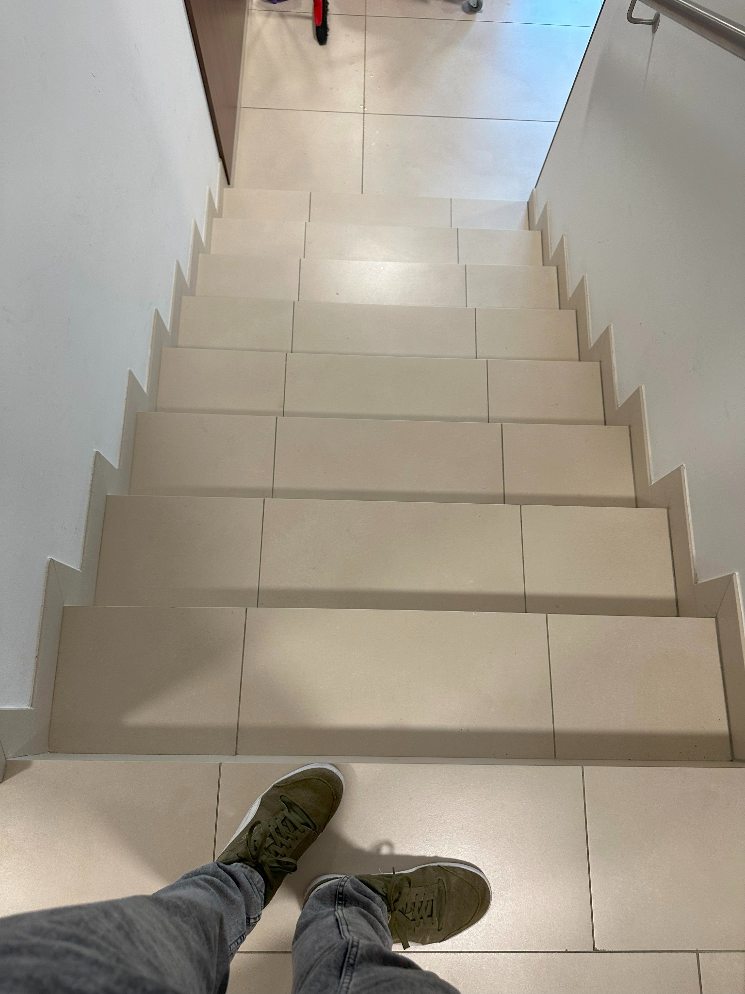 Schody wyłożone jasnymi, kwadratowymi płytkami, widok z góry na stopy w zielonych butach sportowych i szarych dżinsach.