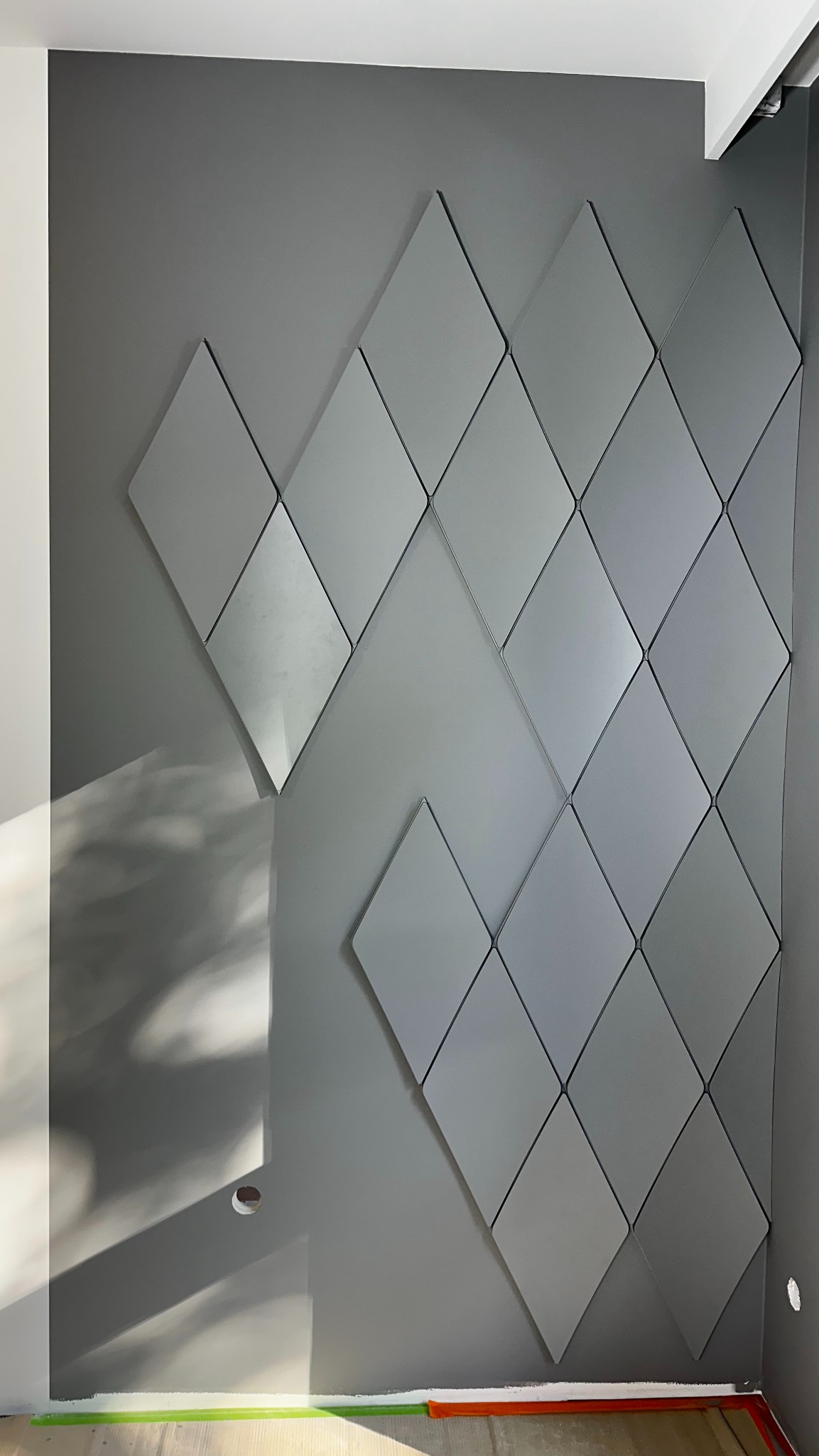 Szara ściana z dekoracyjnymi rombami, widoczny otwór na gniazdko elektryczne i zabezpieczenie podłogi taśmą malarską.