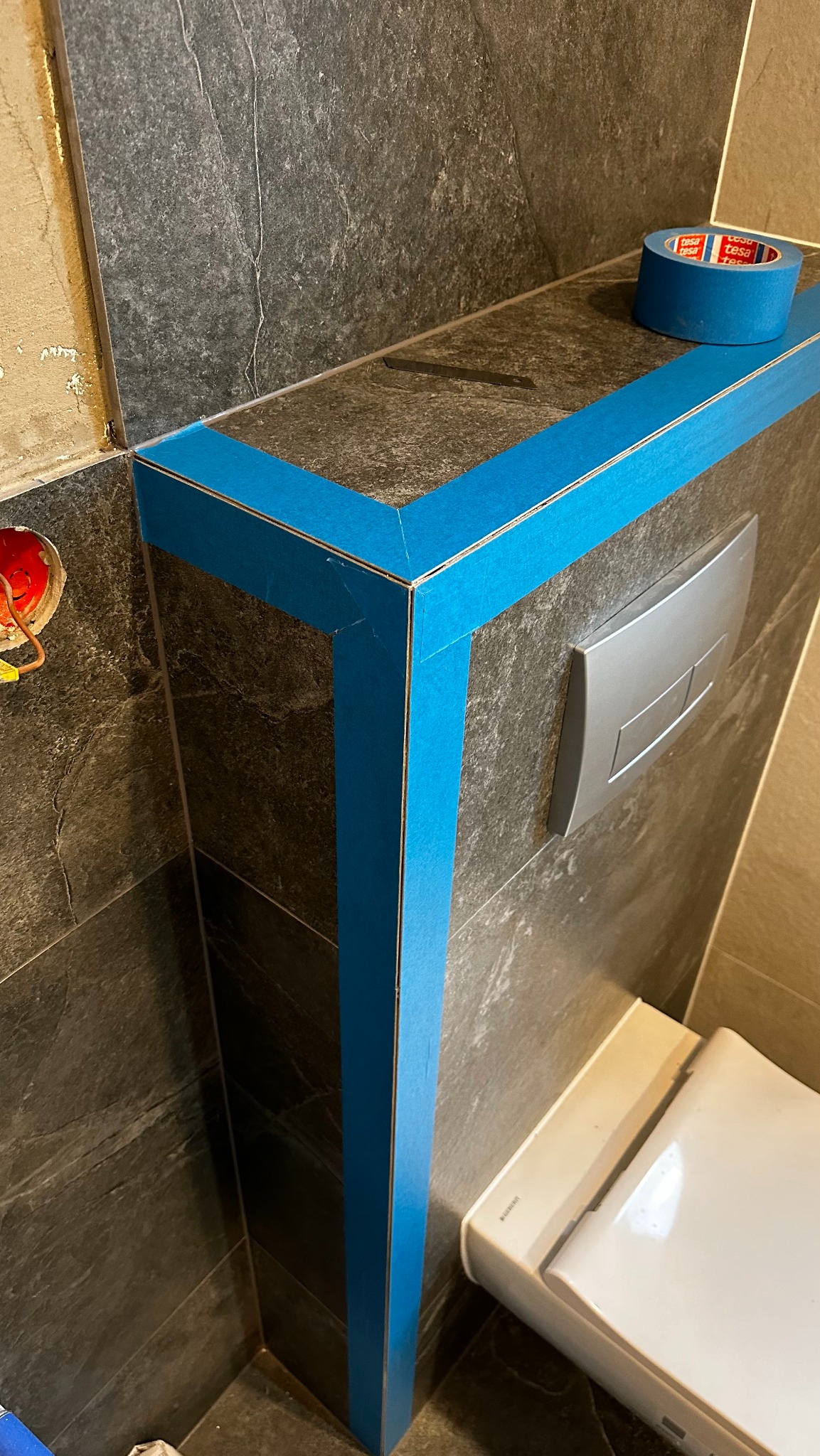 Ciemnoszara okładzina ściany z zamontowanym stelażem podtynkowym WC, zabezpieczona niebieską taśmą malarską Tesa przed fugowaniem, widoczna rolka taśmy i fragment toalety.