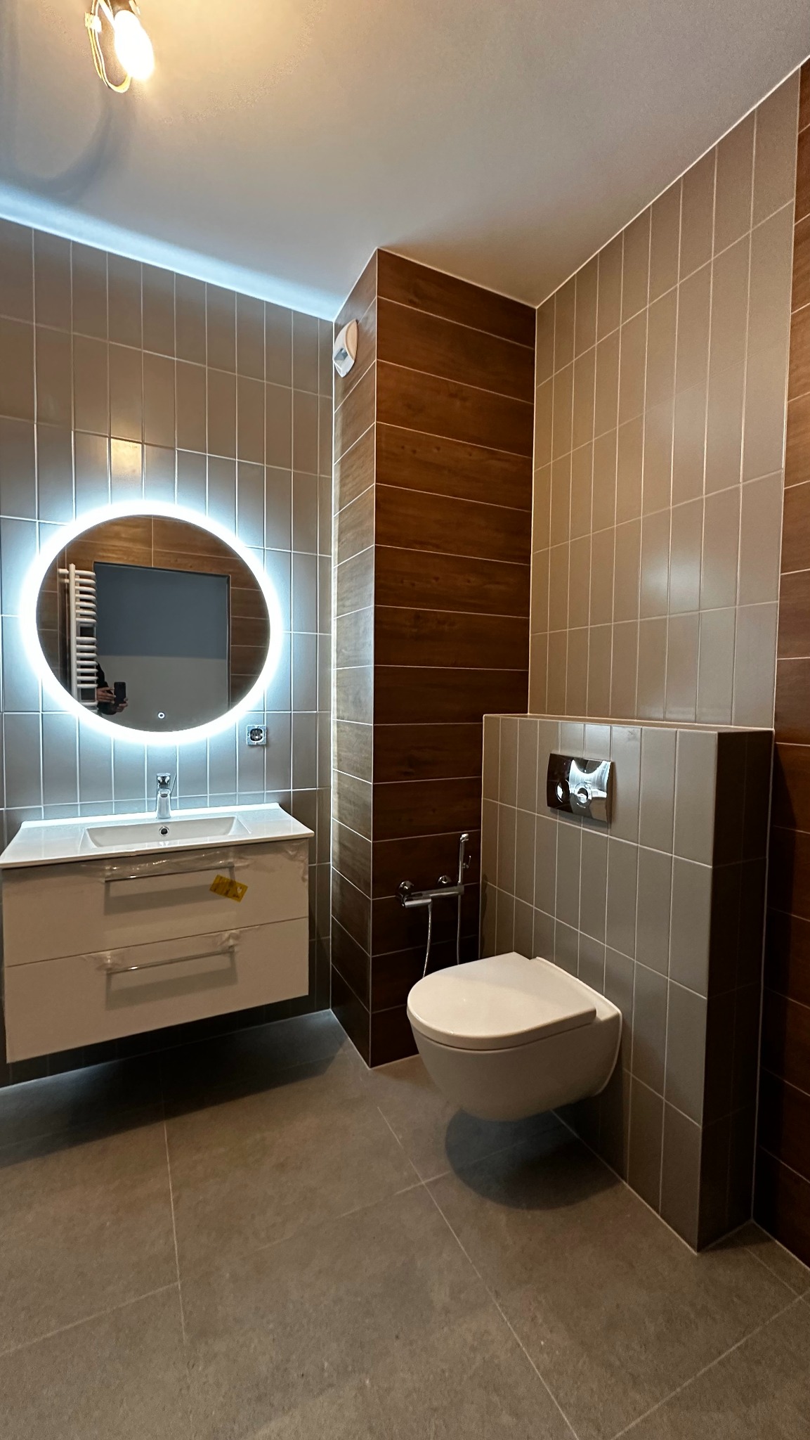 Nowoczesna łazienka z podwieszaną toaletą, umywalką z szafką i okrągłym lustrem z podświetleniem LED. Brązowe i szare płytki na ścianach. Widoczny bidetta.