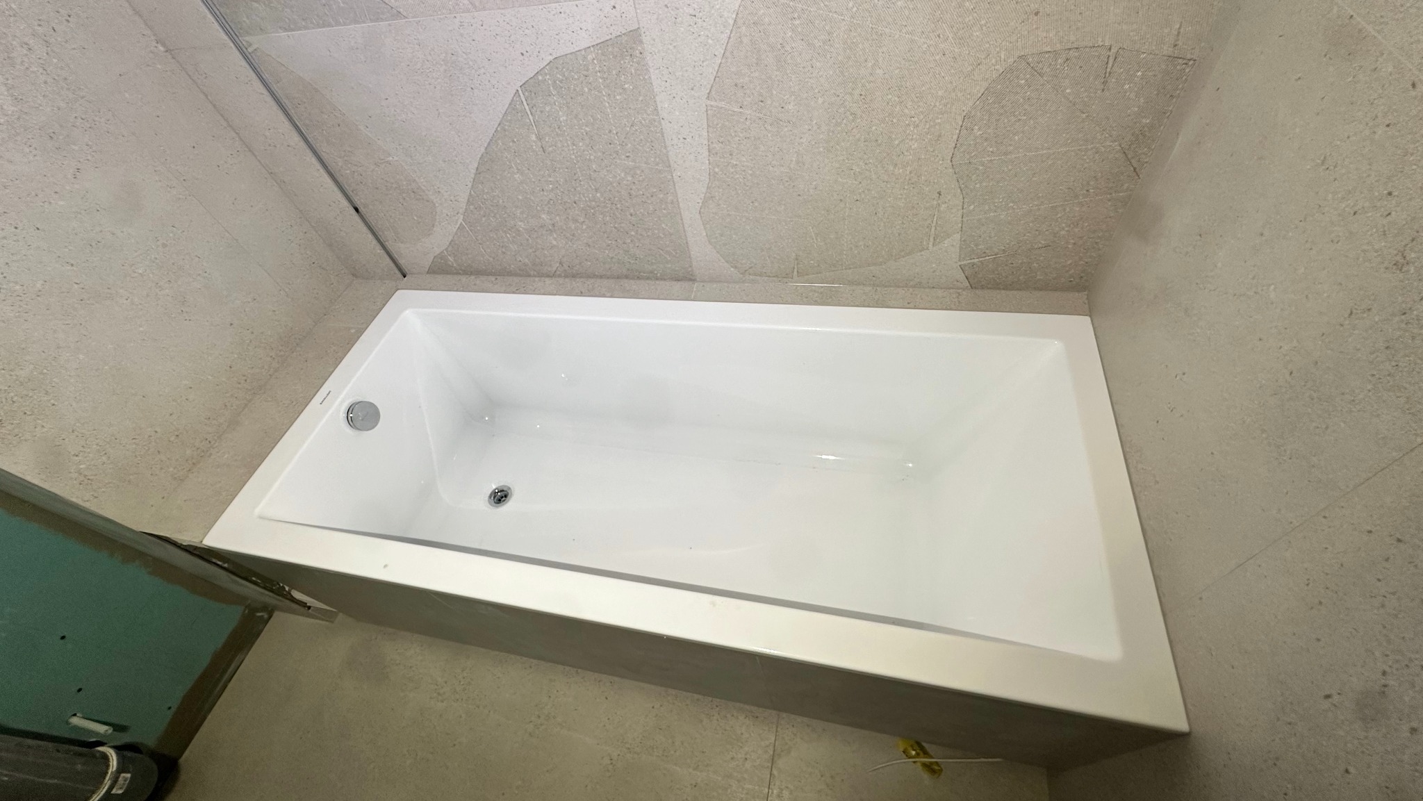 Nowoczesna, biała wanna w obudowie z szarej mozaiki, zamontowana w łazience z płytkami imitującymi kamień w różnych odcieniach szarości, widok z góry, fragment ściany z zieloną płytą...