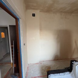 Patryk - Pokój w trakcie przygotowania ścian pod malowanie, widoczne ślady po szpachlowaniu, taśmy malarskie zabezpieczające ościeżnice i listwy przypodłogowe, oraz fragment mebla przykrytego białym...