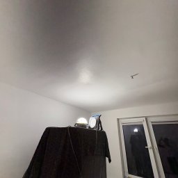 Patryk - Przygotowania do prac wykończeniowych: lampa oświetleniowa na stole przykrytym czarną folią, widoczny fragment sufitu z nierównomierną powierzchnią, okno z zabezpieczoną folią ramą.