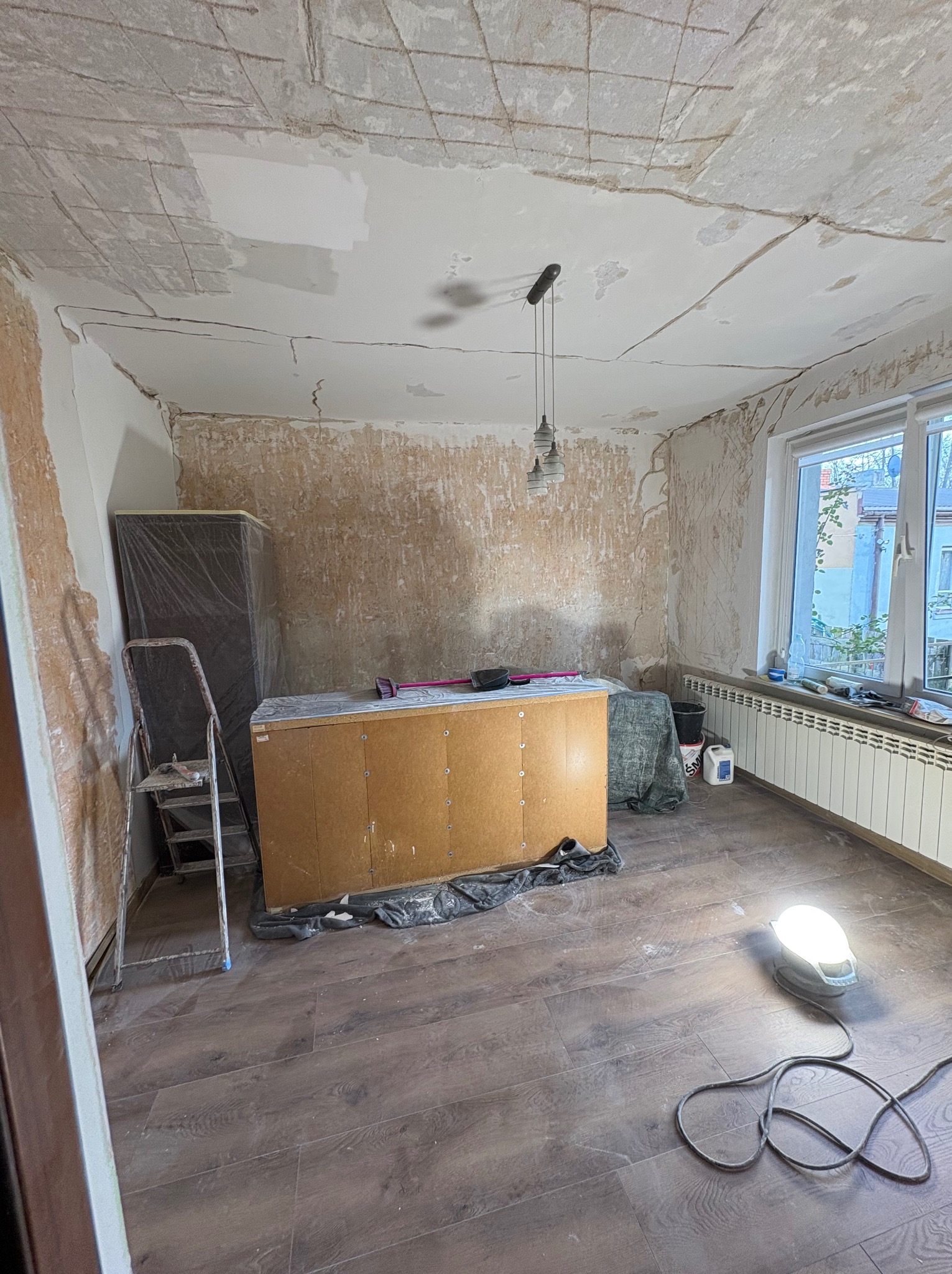 Pokój w trakcie remontu: ściany przygotowane do prac wykończeniowych, widoczne ślady po usuniętej warstwie, sufit z widoczną siatką wzmacniającą, podłoga częściowo zakryta folią ochronną, lampa...