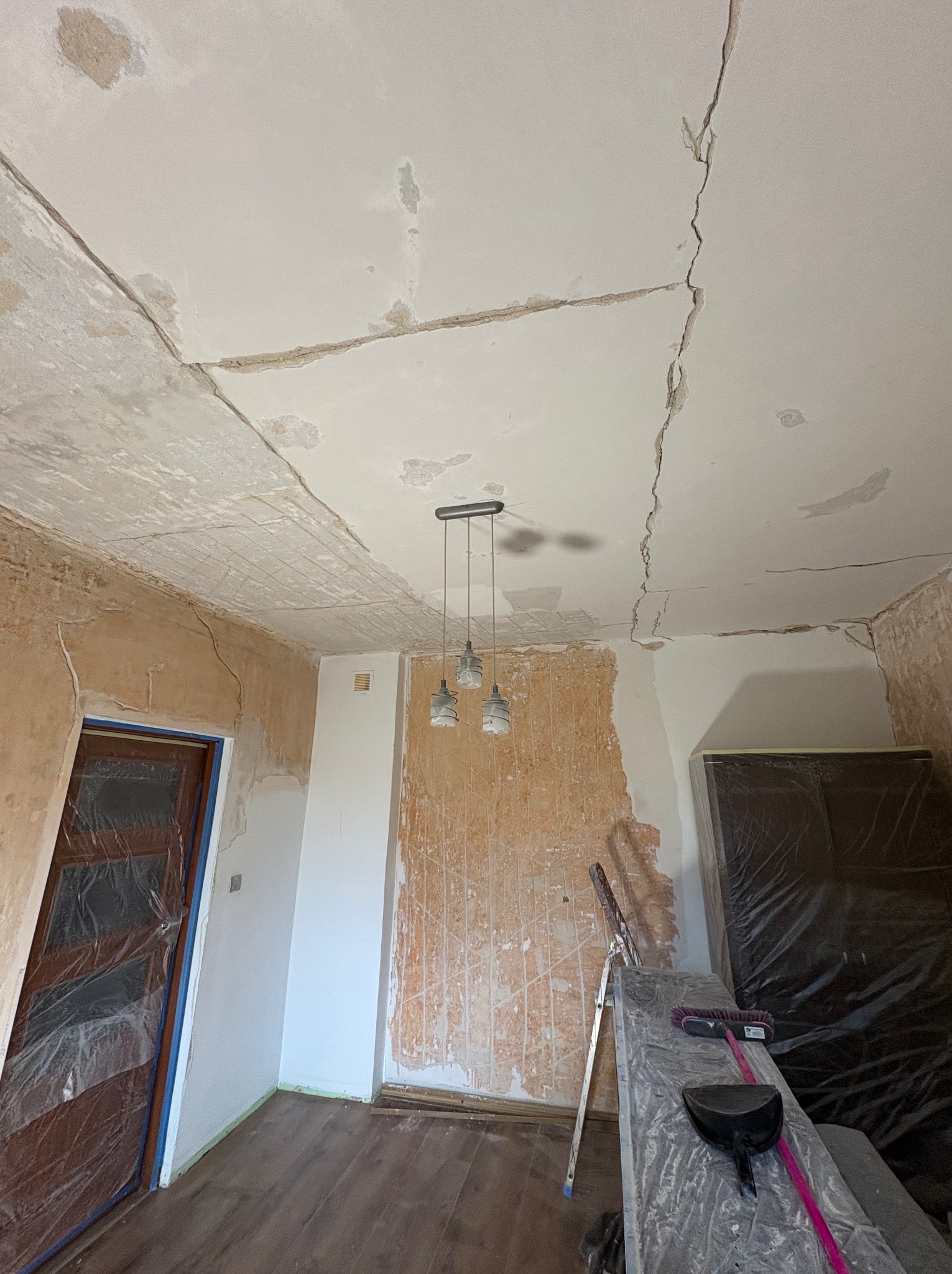 Przygotowanie do remontu: Pokój z pęknięciami na suficie, ściany z resztkami starej farby, zabezpieczone drzwi folią, drabina i narzędzia w rogu.