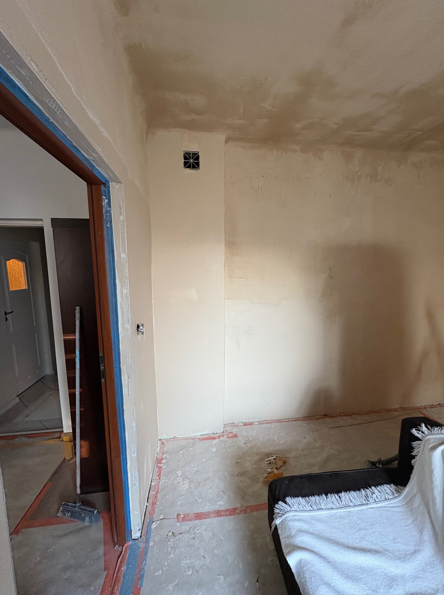 Pokój w trakcie przygotowania ścian pod malowanie, widoczne ślady po szpachlowaniu, taśmy malarskie zabezpieczające ościeżnice i listwy przypodłogowe, oraz fragment mebla przykrytego białym...