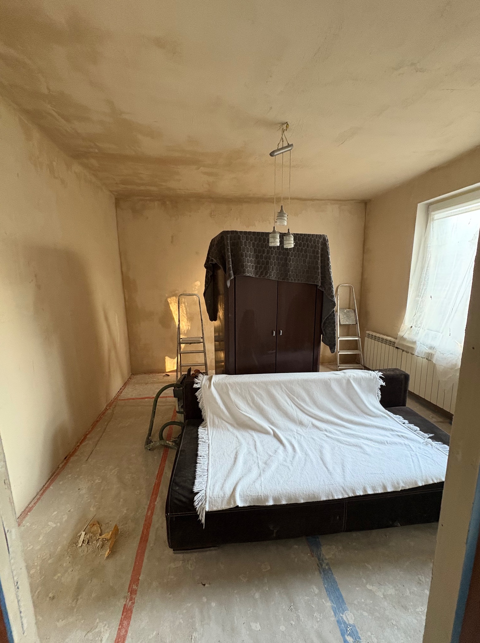 Pokój w trakcie szpachlowania: ściany pokryte świeżo nałożoną, nierówną warstwą szpachli, zabezpieczone meble pod przykryciem, widoczne ślady taśmy malarskiej na podłodze i drabina oparta o szafę.