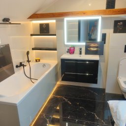 AD RenoHome - Nowoczesna łazienka na poddaszu z czarną marmurową podłogą z złotymi żyłami, białą wanną z czarną armaturą, czarną szafką pod umywalką i podświetlanym lustrem, akcentowana drewnianą belką stropową...