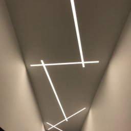 AD RenoHome - Nowoczesne oświetlenie sufitowe z liniowymi lampami LED ułożonymi pod kątem, tworzącymi geometryczny wzór na gładkim, jasnym suficie.