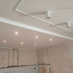 AD RenoHome - Wnętrze w trakcie remontu: biały sufit z dekoracyjną sztukaterią i punktowymi oprawami oświetleniowymi, ściany częściowo pokryte płytkami i zaprawą, widoczna drabina. Na ścianach widoczne notatki...