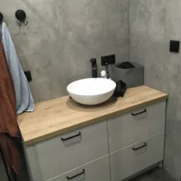 Nowoczesna łazienka z szarą płytką, białą umywalką nablatową i szafką z drewnianym blatem, uzupełniona o czarne detale: bateria, uchwyty, haczyki i akcesoria.