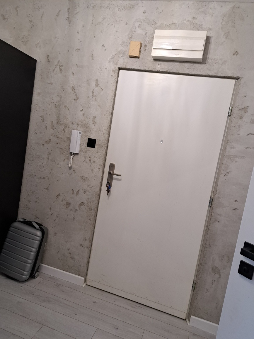 Wejście do mieszkania z białą, minimalistyczną ścianą o teksturze betonu, białe drzwi z wizjerem i kluczem w zamku, szara walizka przy ścianie, domofon i skrzynka elektryczna nad drzwiami.