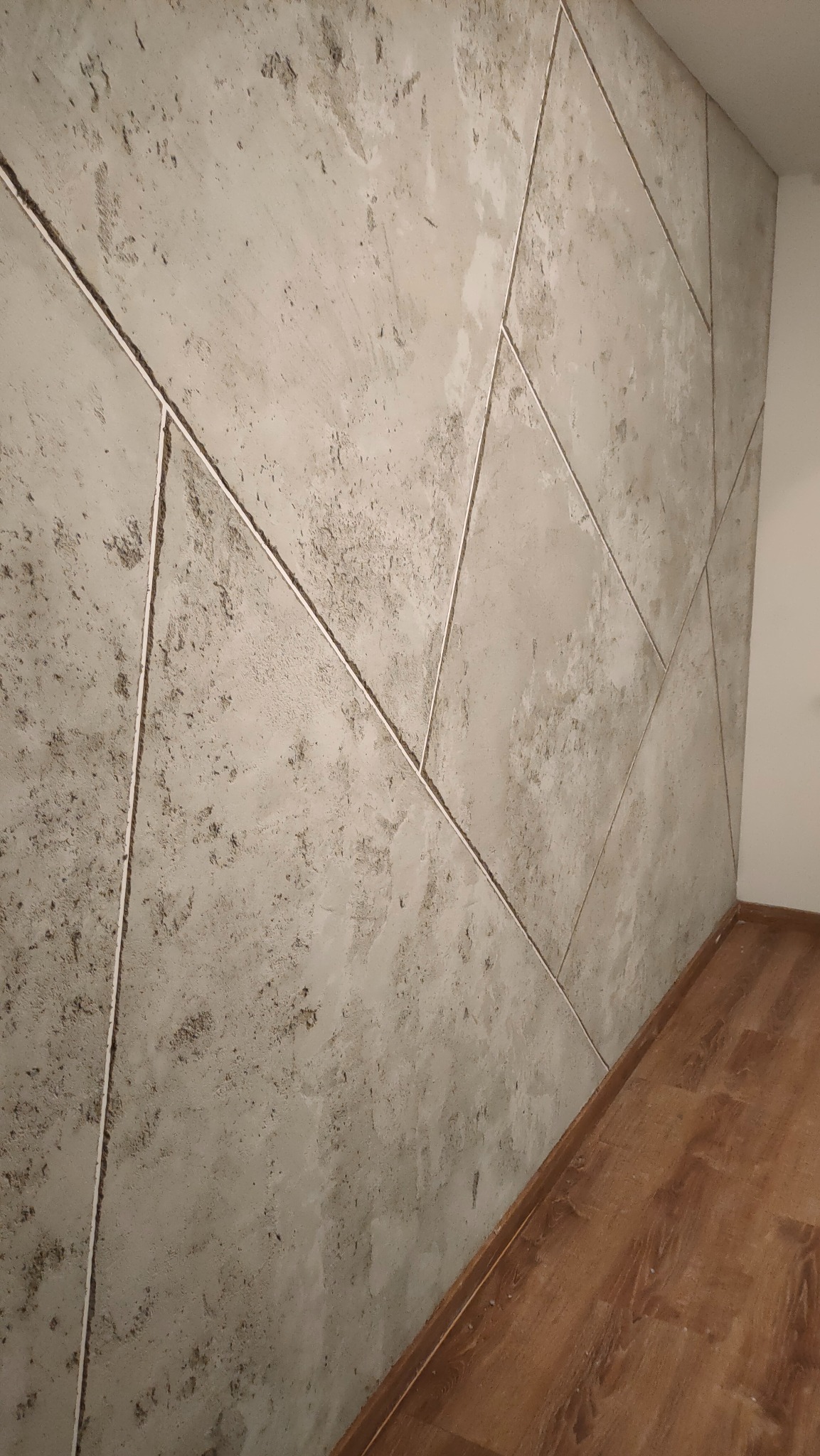 Dekoracyjna ściana z szarego betonu architektonicznego, podzielona na geometryczne panele z delikatnymi wżerami, widoczna pod kątem, z fragmentem drewnianej podłogi i białym narożnikiem ściany.