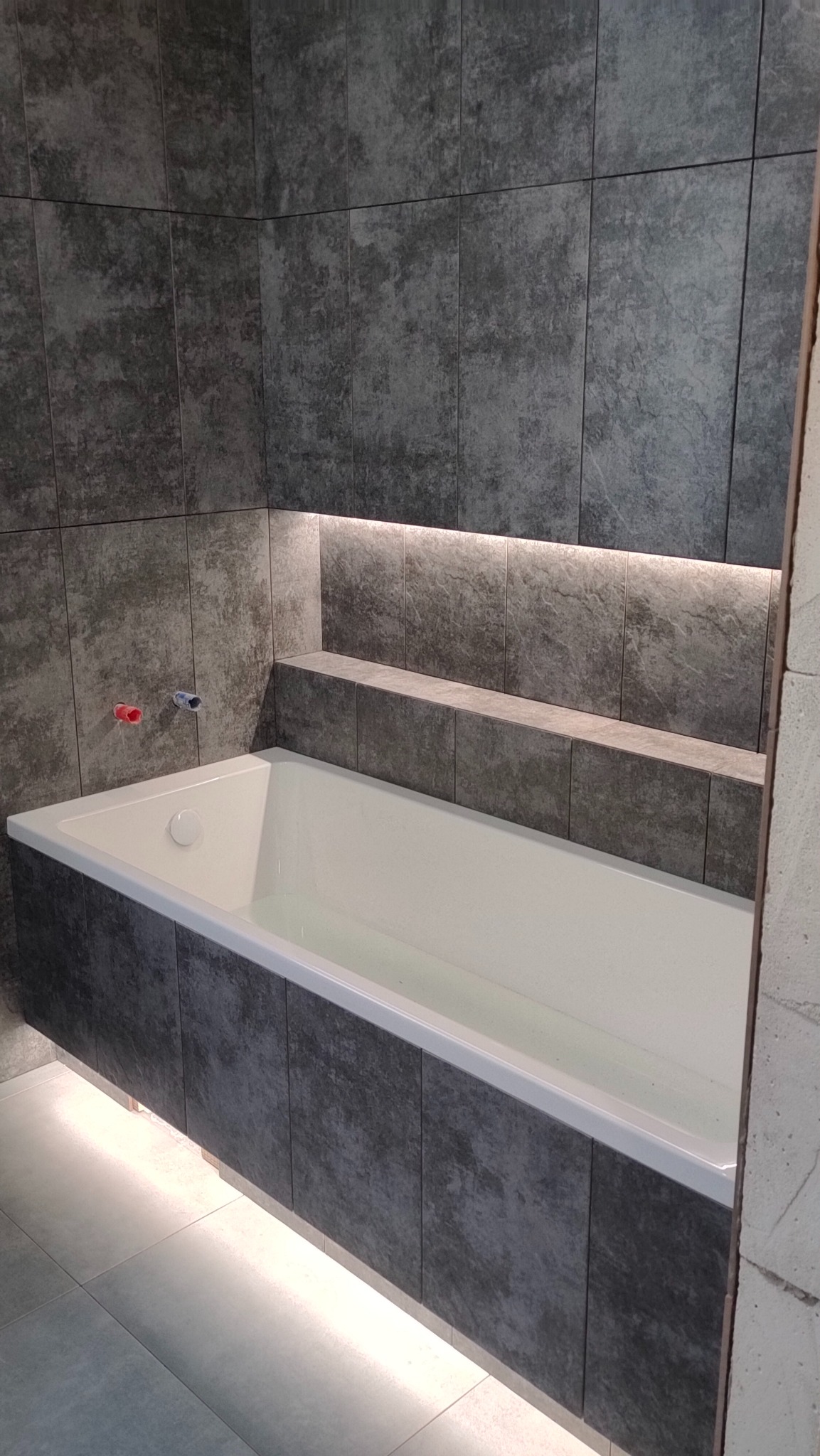 Nowoczesna łazienka z wanną obłożoną ciemnymi płytkami imitującymi beton, podświetlenie LED pod wanną i półką, minimalistyczna armatura.