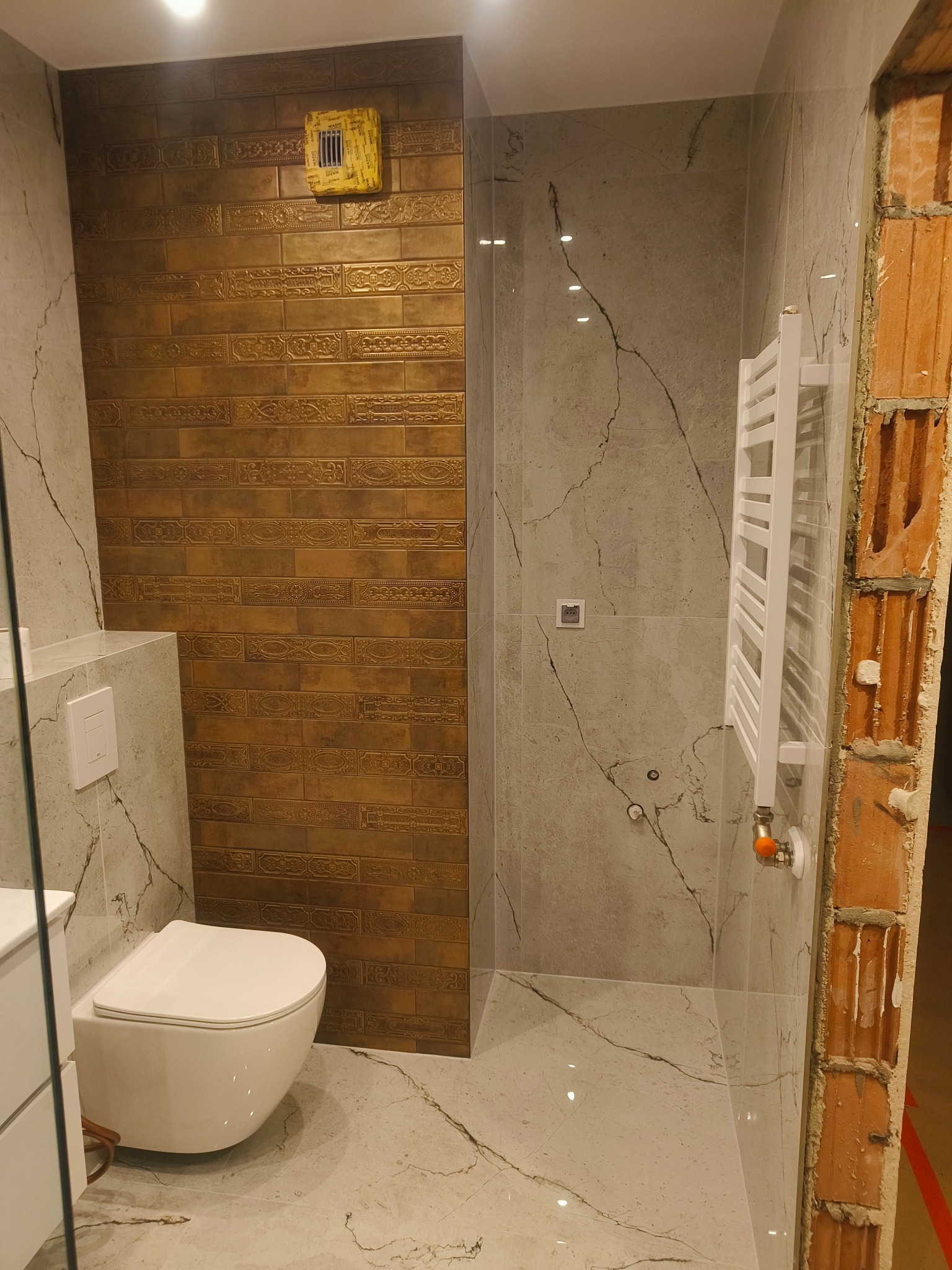 Nowoczesna łazienka z wiszącą toaletą, grzejnikiem drabinkowym, złotymi płytkami dekoracyjnymi i marmurowymi płytami, jeszcze w trakcie remontu z widocznymi surowymi cegłami.