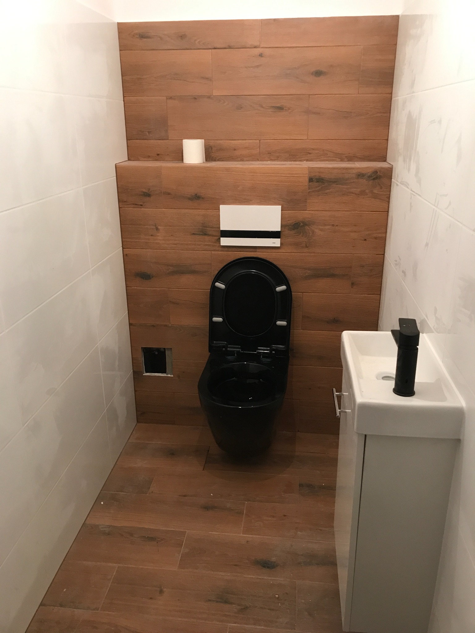 Nowoczesna toaleta w małej łazience z białymi płytkami i drewnopodobnymi panelami, wisząca czarna muszla klozetowa i mała biała umywalka z czarną baterią.