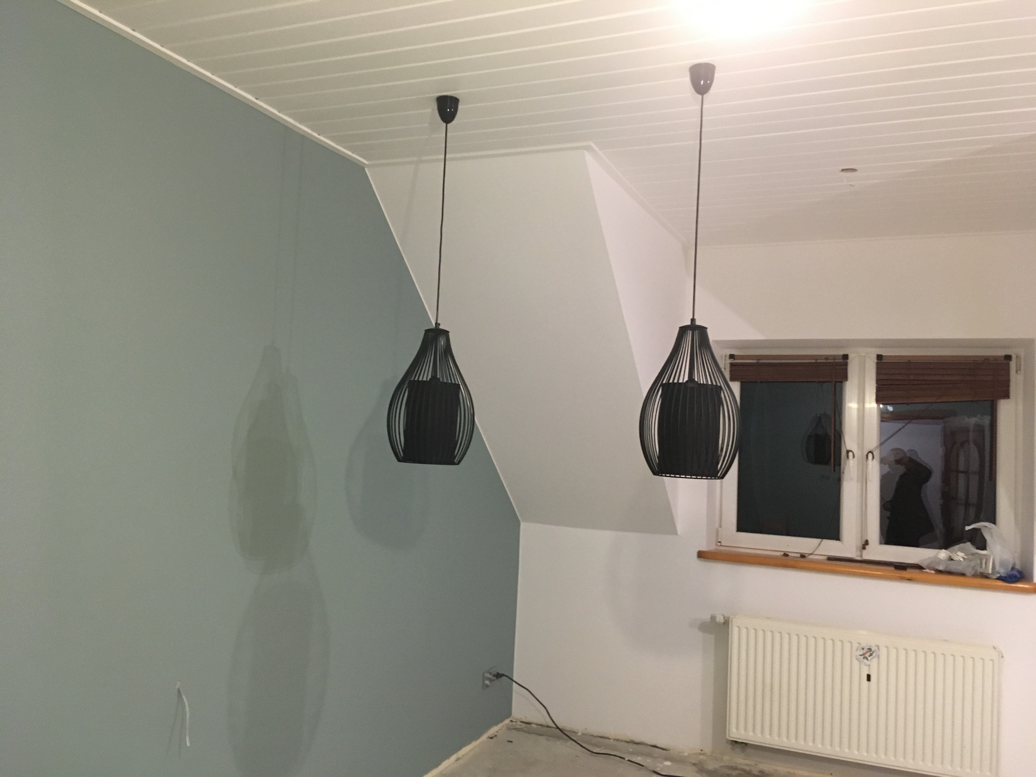 Świeżo pomalowane na szaro ściany w pokoju ze skośnym sufitem, widoczne dwie czarne lampy wiszące, biały grzejnik pod oknem i surowa podłoga przed wykończeniem.