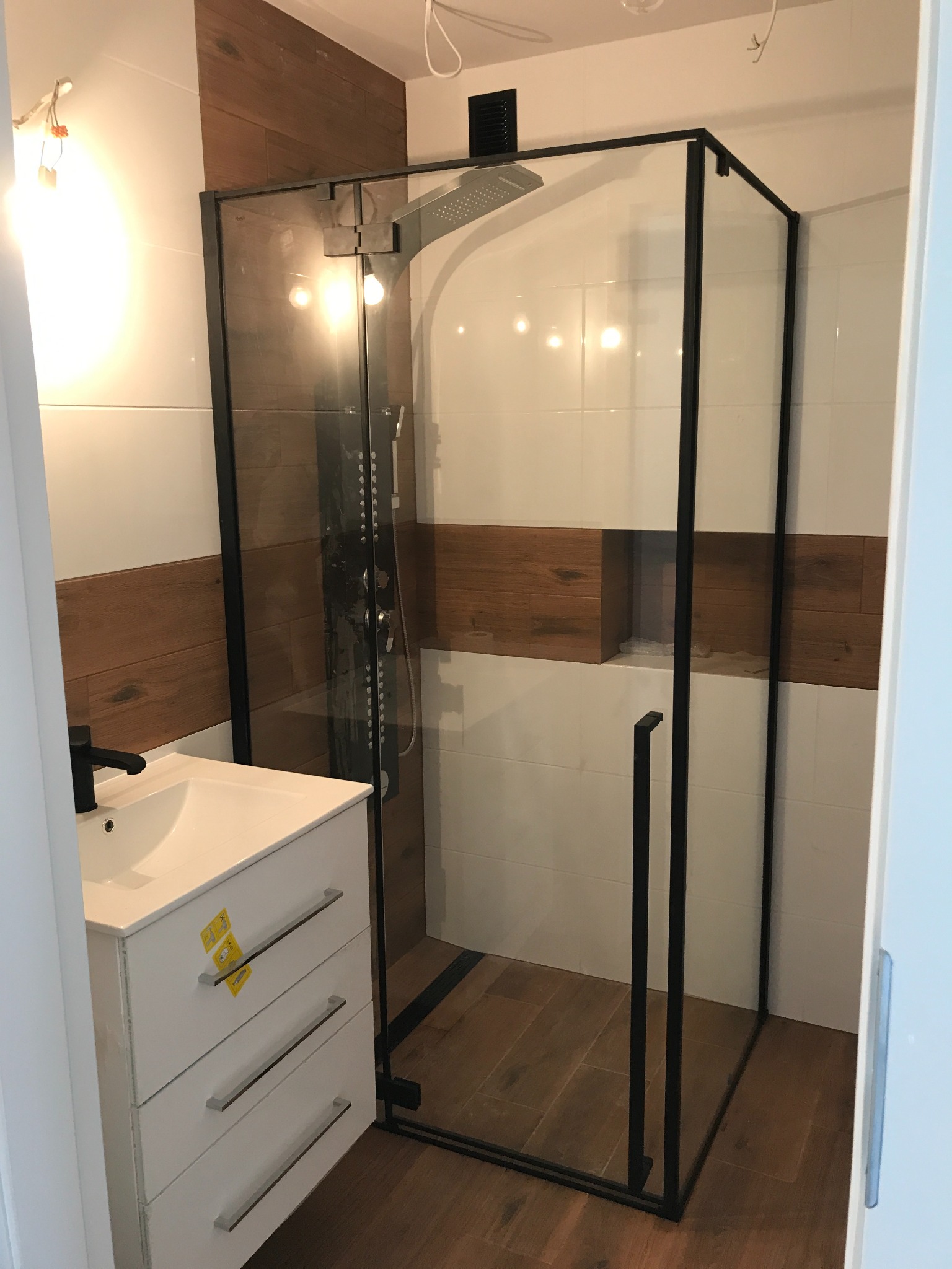 Nowoczesna łazienka z kabiną prysznicową typu walk-in z czarnymi profilami, białą umywalką nablatową z czarną armaturą i płytkami imitującymi drewno na ścianach.