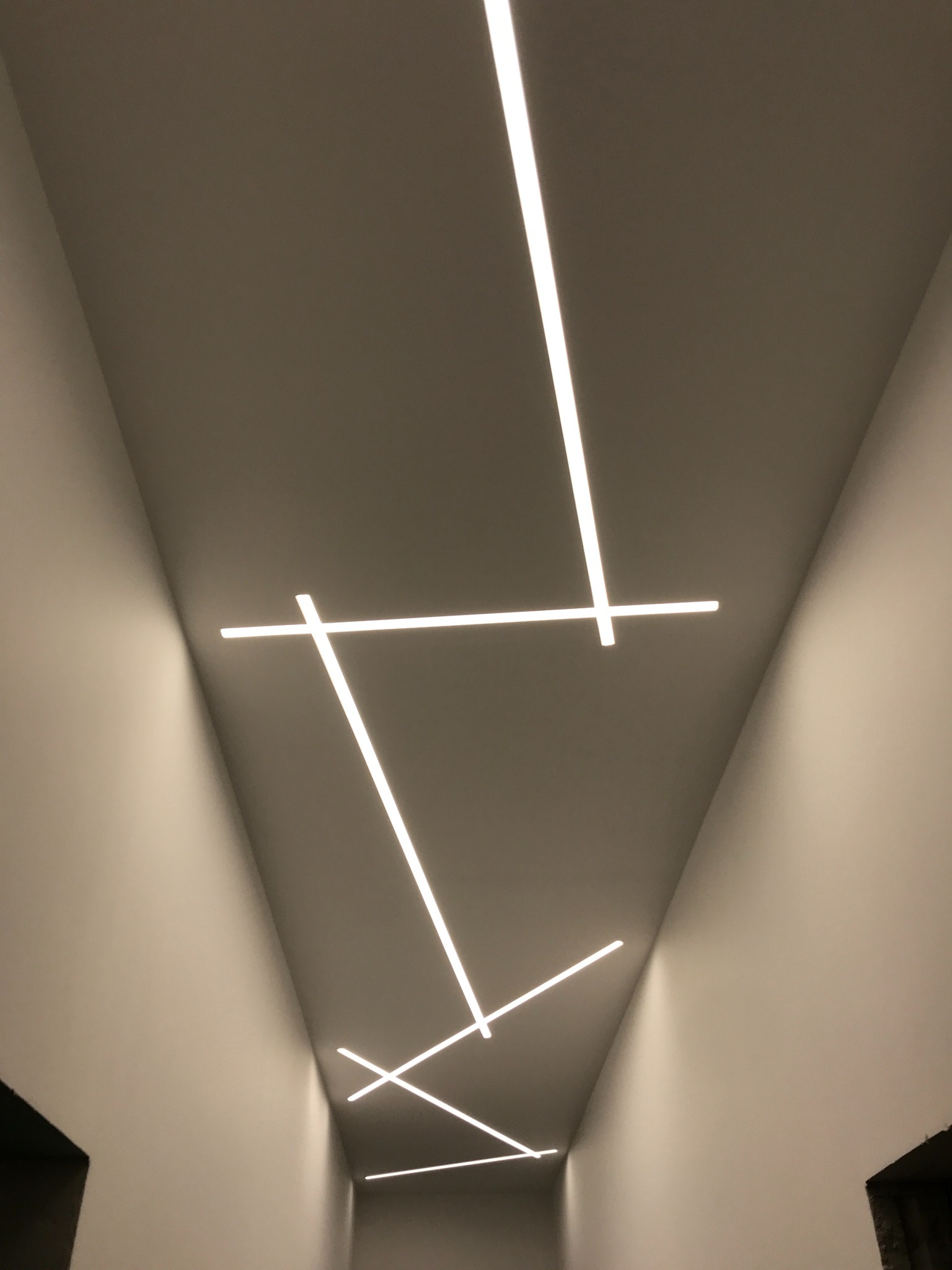 Nowoczesne oświetlenie sufitowe z liniowymi lampami LED ułożonymi pod kątem, tworzącymi geometryczny wzór na gładkim, jasnym suficie.