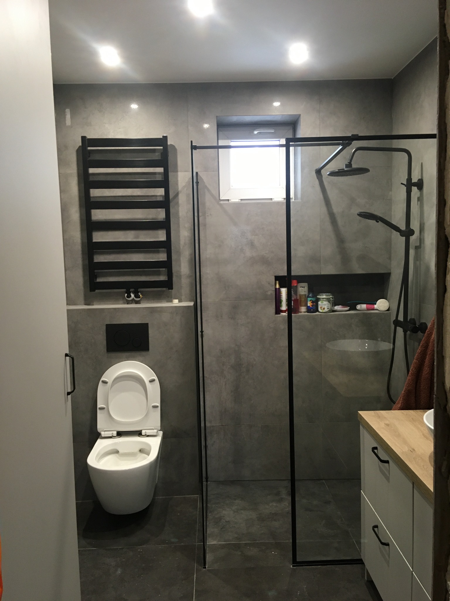 Nowoczesna łazienka w odcieniach szarości z czarnymi akcentami: kabina prysznicowa typu walk-in, wisząca toaleta, grzejnik drabinkowy i szafka z drewnianym blatem.