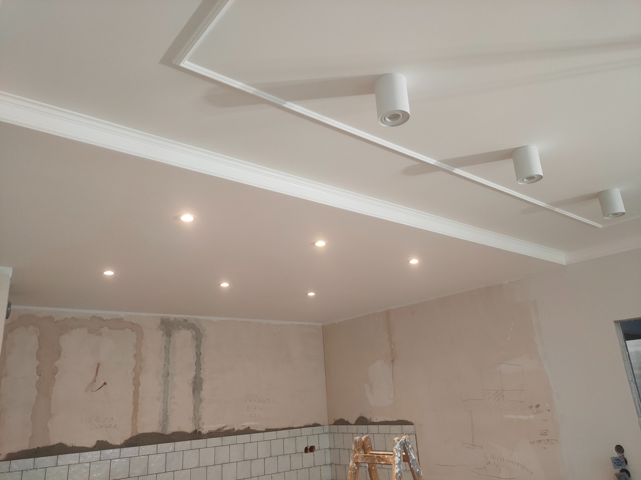 Wnętrze w trakcie remontu: biały sufit z dekoracyjną sztukaterią i punktowymi oprawami oświetleniowymi, ściany częściowo pokryte płytkami i zaprawą, widoczna drabina. Na ścianach widoczne notatki...