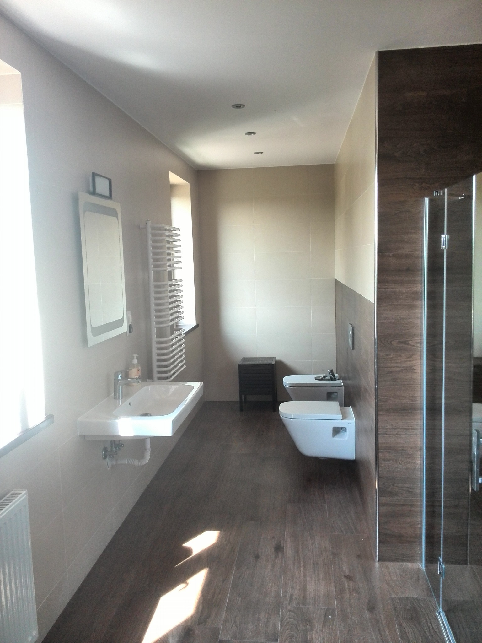 Nowoczesna łazienka z podwieszaną toaletą i bidetem, umywalką nablatową, grzejnikiem drabinkowym i kabiną prysznicową z brązowymi płytkami imitującymi drewno na ścianie.