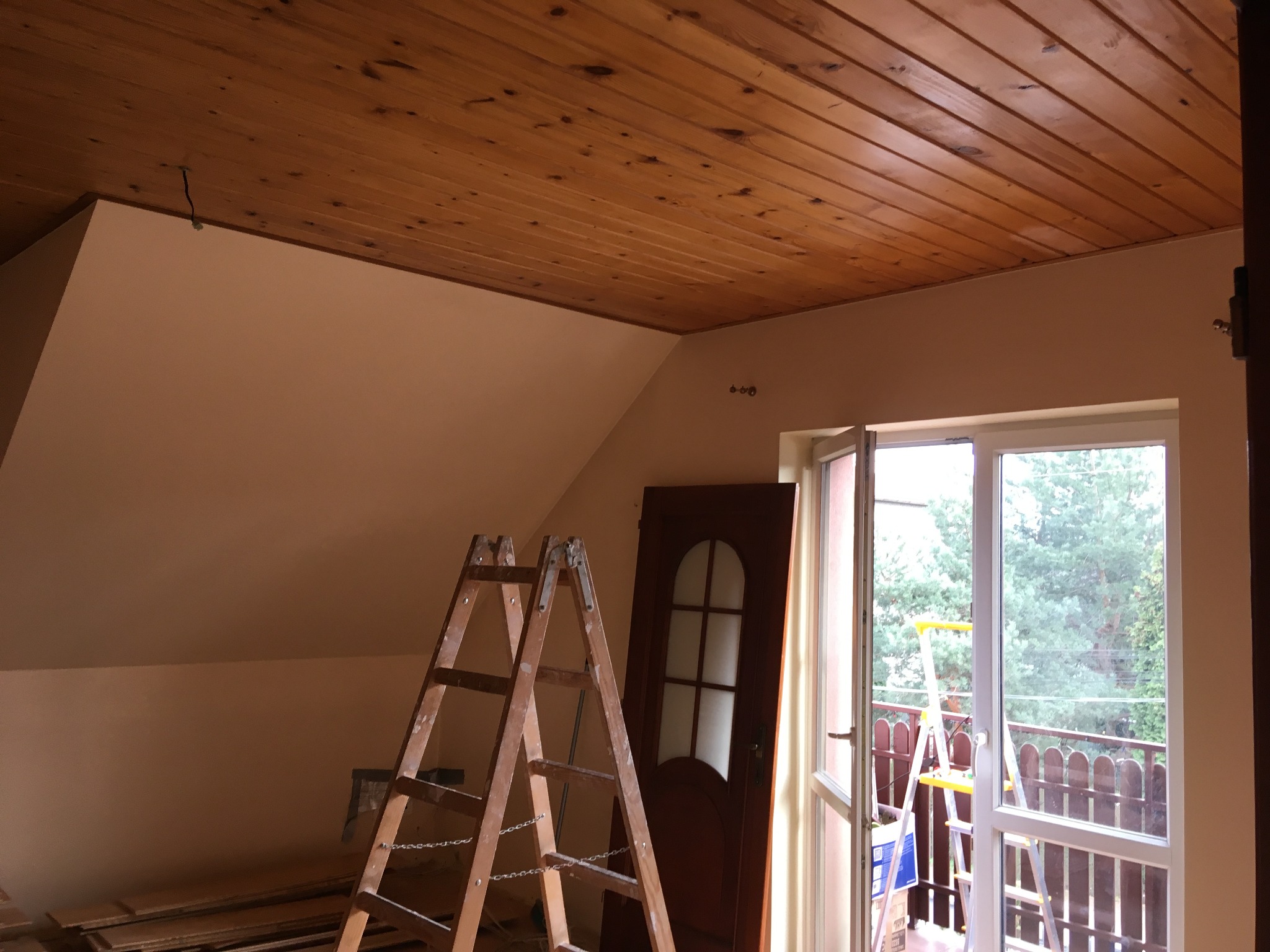 Poddasze w trakcie prac wykończeniowych: drewniana boazeria na suficie, jasne ściany, drabina malarska, drzwi wewnętrzne i okno balkonowe z widokiem na zieleń.