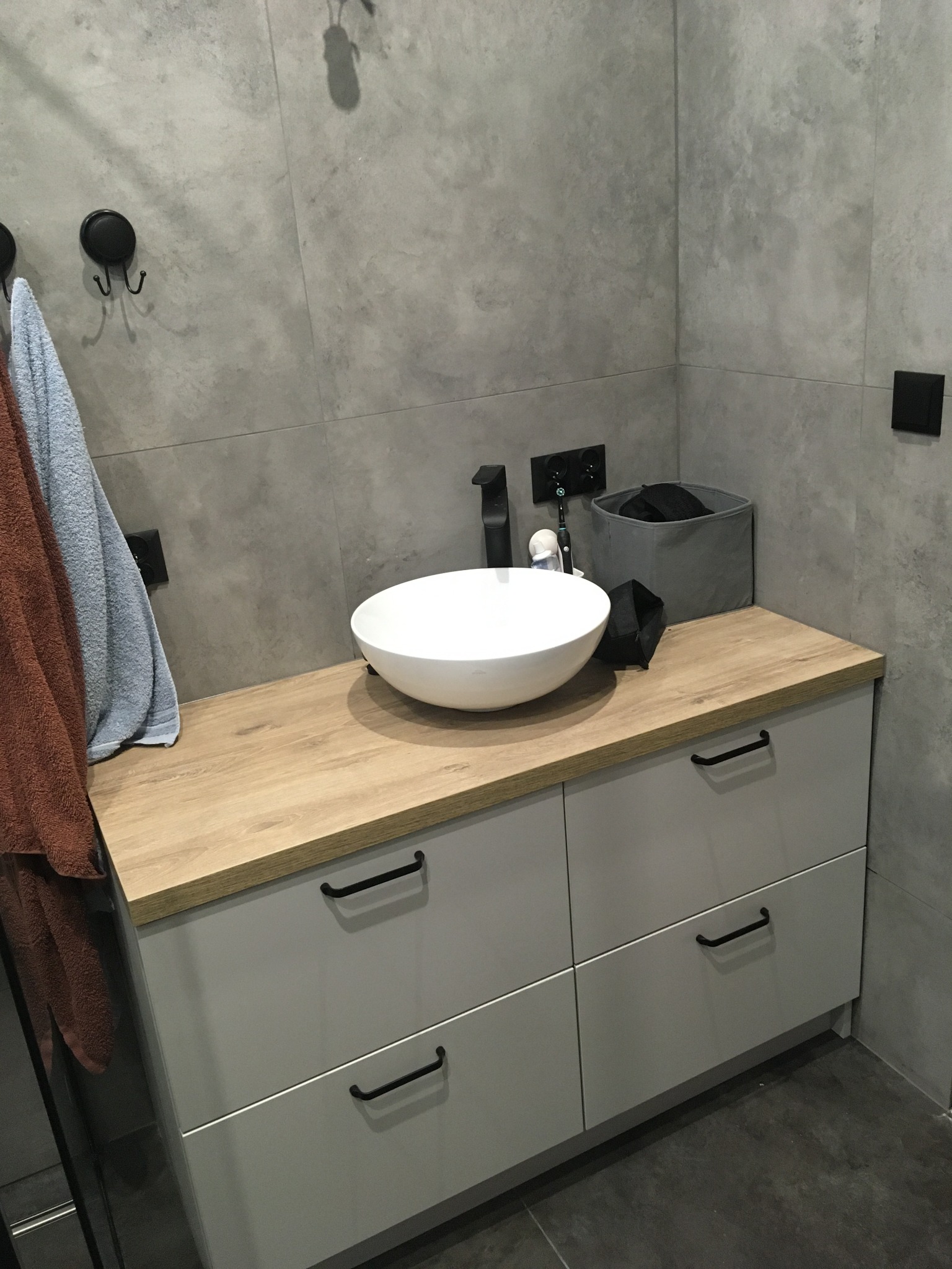 Nowoczesna łazienka z szarą płytką, białą umywalką nablatową i szafką z drewnianym blatem, uzupełniona o czarne detale: bateria, uchwyty, haczyki i akcesoria.