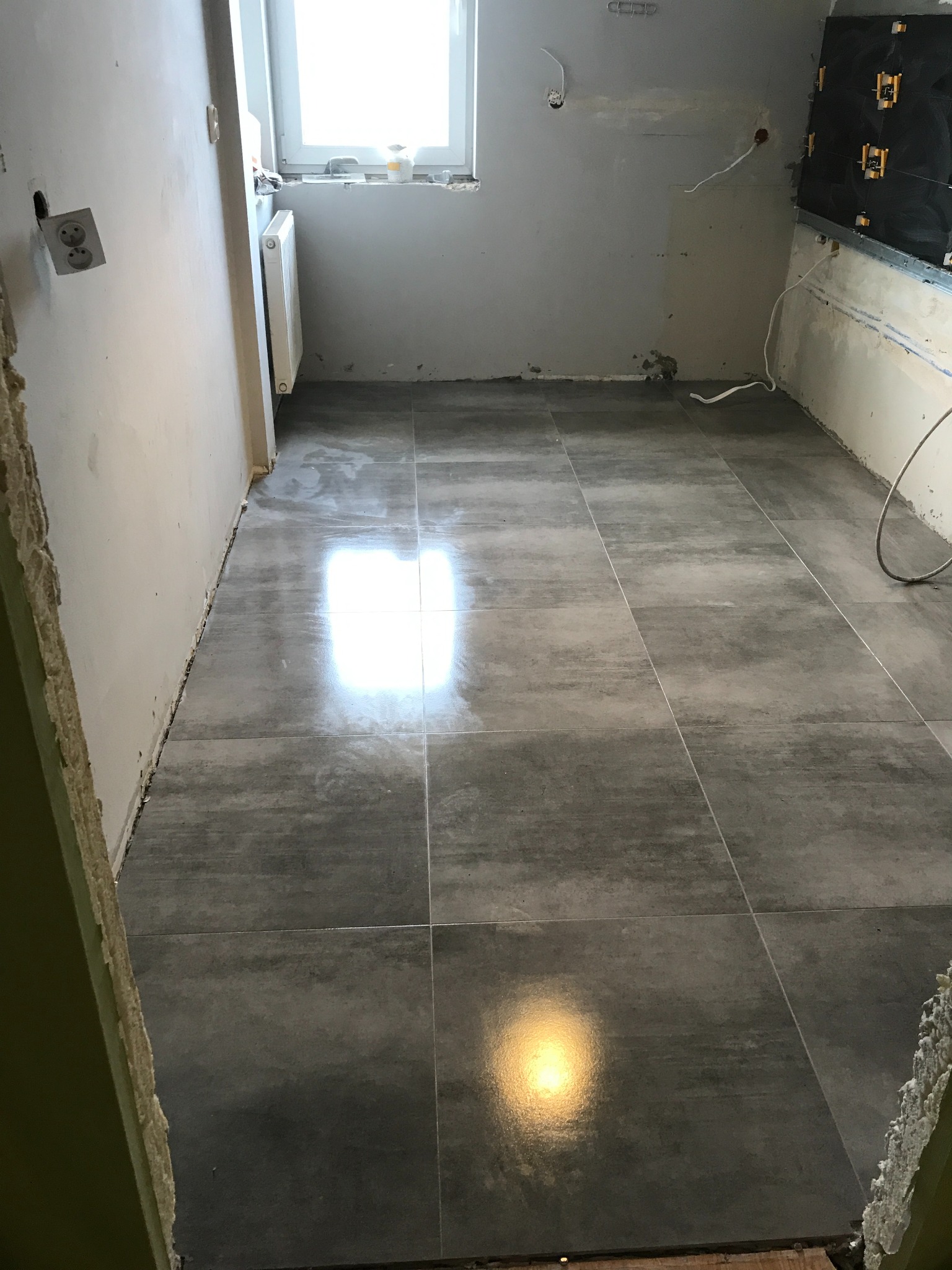 Szare, matowe płytki podłogowe ułożone w trakcie remontu łazienki, widoczne niedokończone ściany i instalacje.