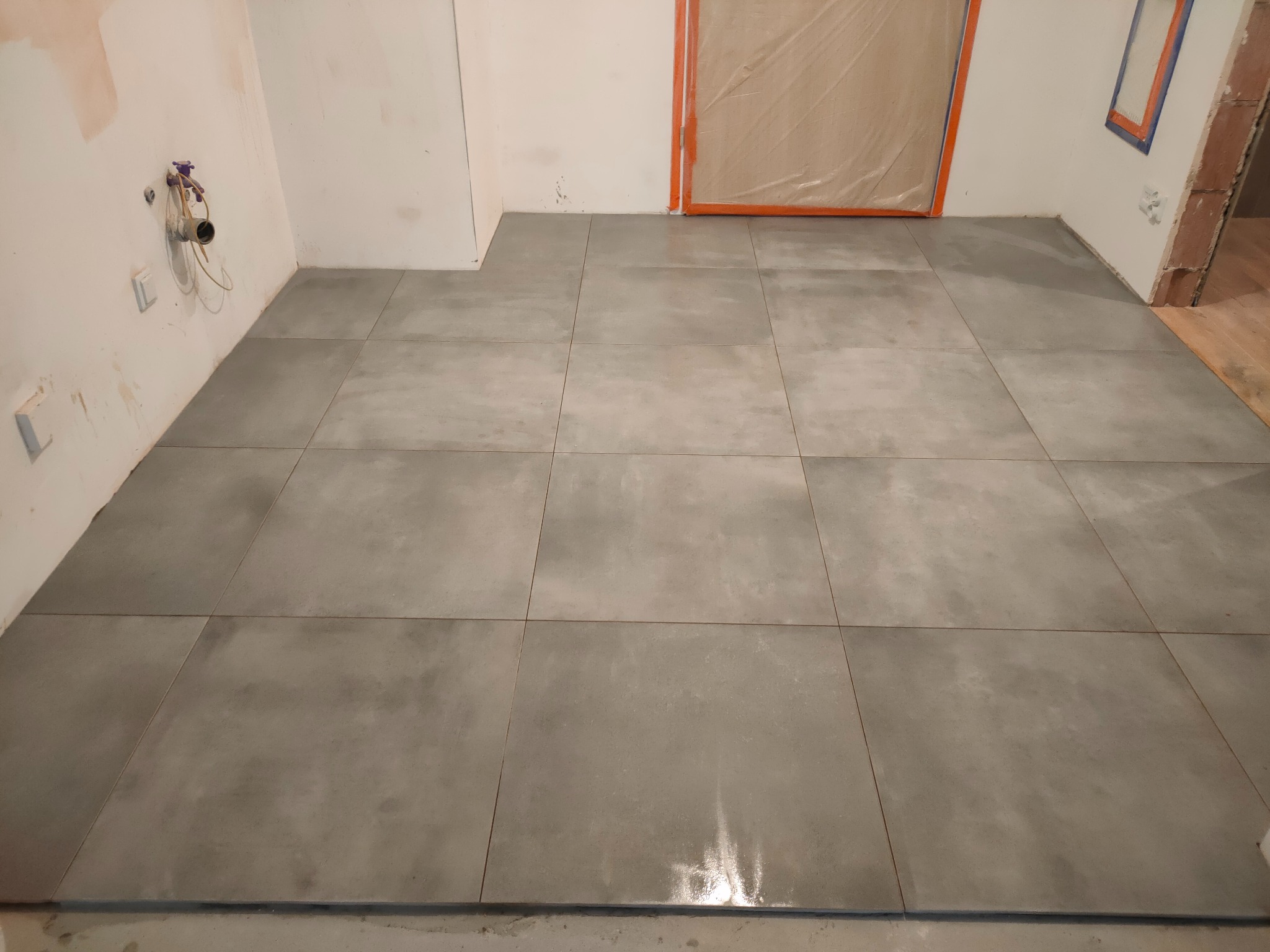 Świeżo położona, błyszcząca, szara podłoga z dużych płytek w trakcie remontu łazienki, widoczne zabezpieczone folią drzwi i wystające rury.