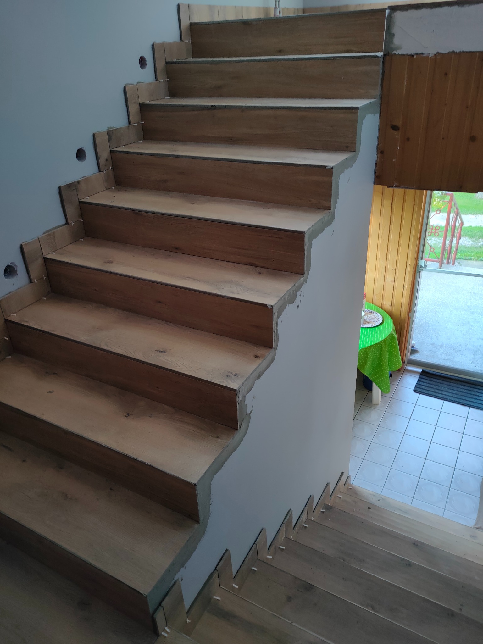 Schody w trakcie układania płytek imitujących drewno, widoczne krzyżyki dystansowe, surowa ściana i wyjście na zewnątrz.