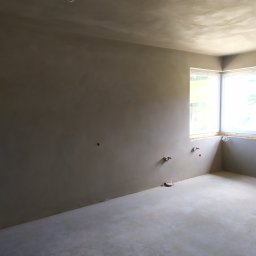 Tynki maszynowe tradycyjne - Surowe wnętrze pokoju po tynkowaniu maszynowym, widoczne przygotowane instalacje elektryczne i hydrauliczne, narożne okno z widokiem na zieleń.