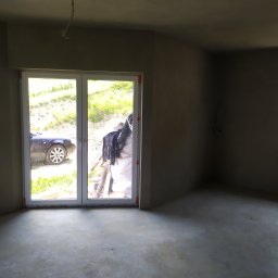 Tynki maszynowe tradycyjne - Surowe wnętrze po nałożeniu tynku, widoczne kable wystające z sufitu, okno z widokiem na samochód i teren zielony.