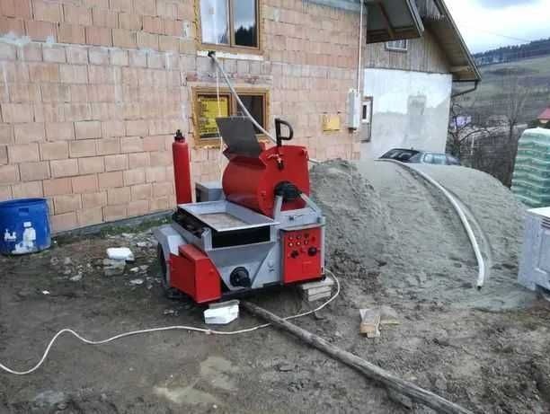 Czerwono-szara maszyna do tynkowania stoi obok sterty piasku na placu budowy, z widocznym fragmentem ceglanego budynku w tle.